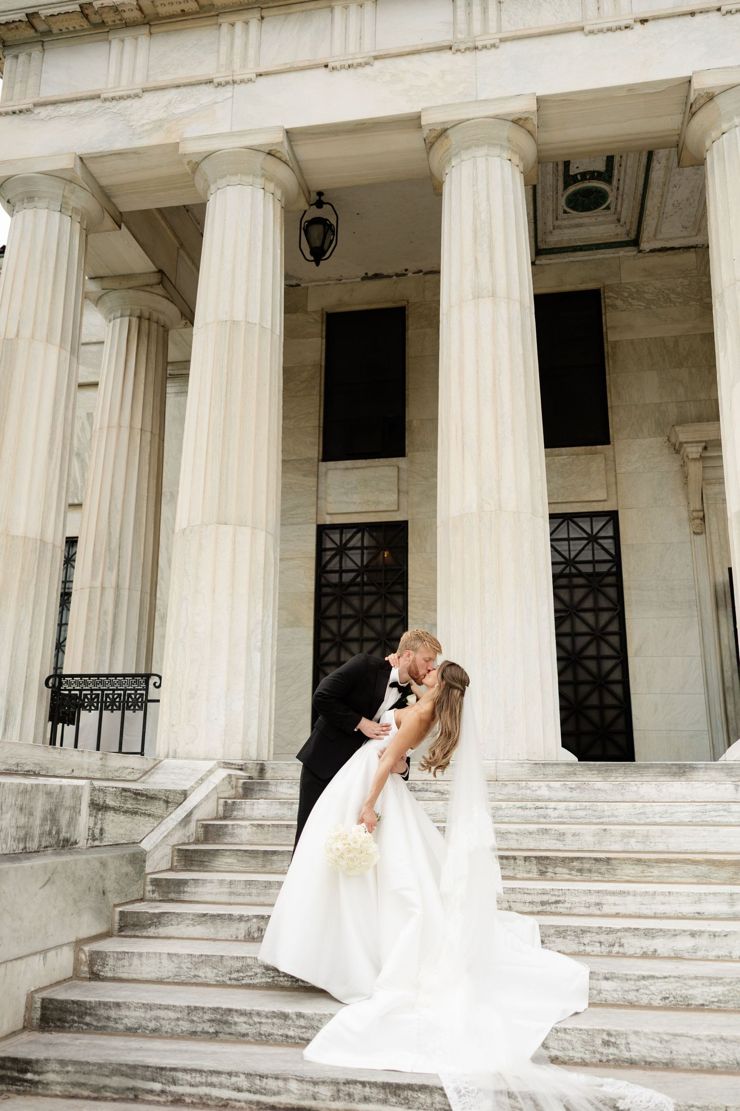 Bride and groom share a romantic kiss on grand marble steps of a classical building with tall white columns.