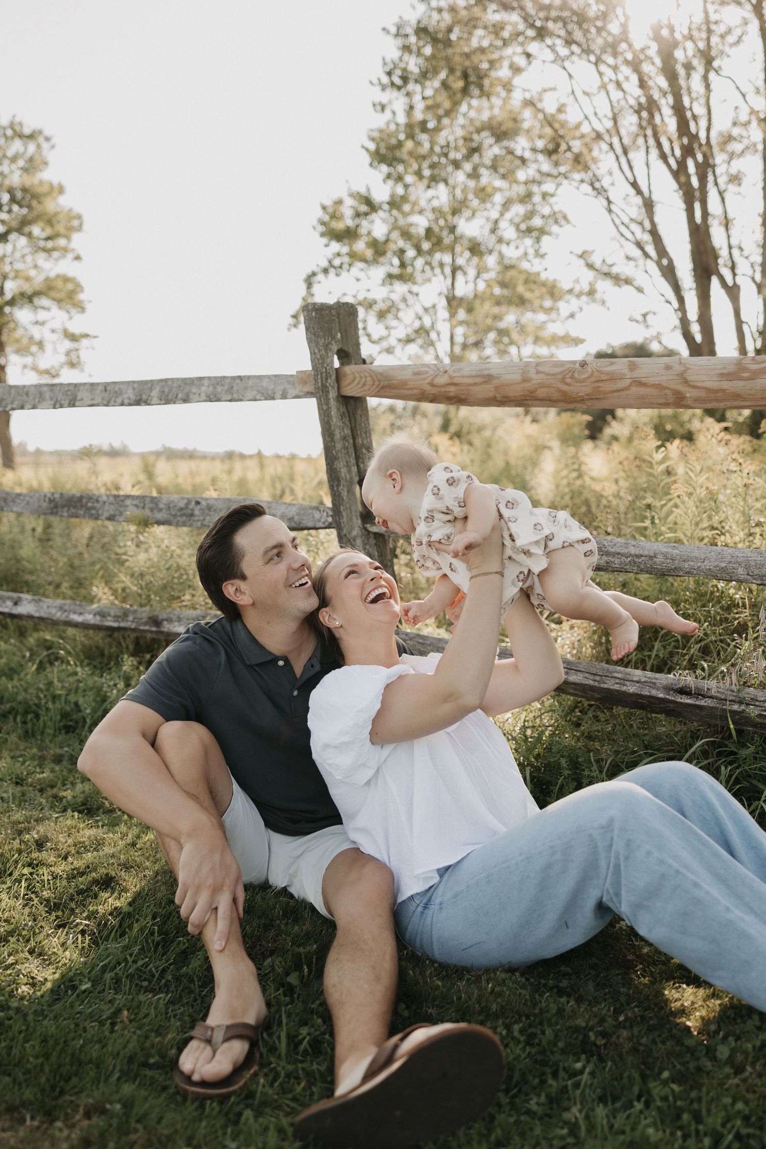 A happy young family sitting on grass by a rustic fence, laughing as mom lifts baby in the air outdoors.