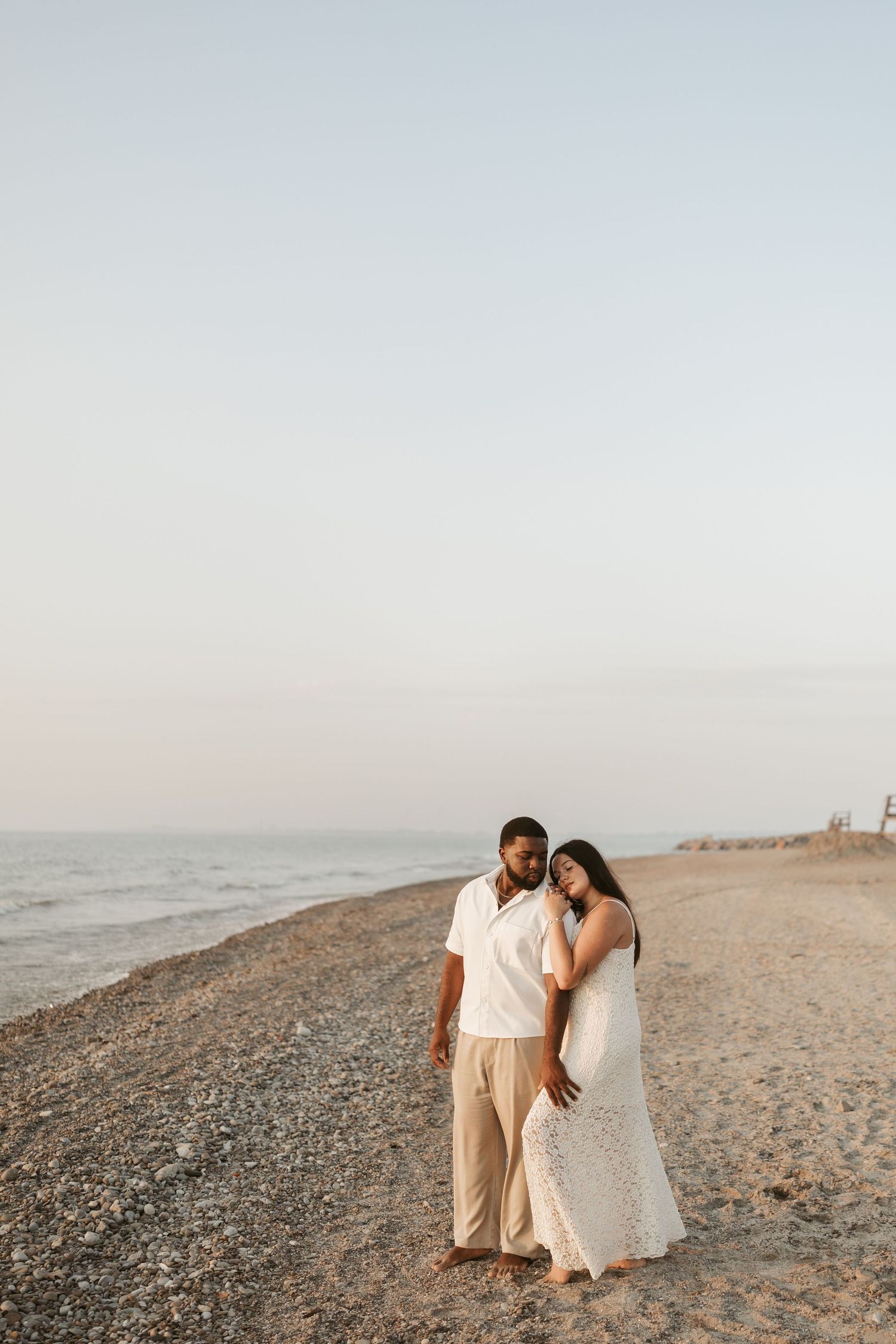A couple walks along a sandy beach at sunset, woman in white lace dress leaning on man in white shirt and tan pants.