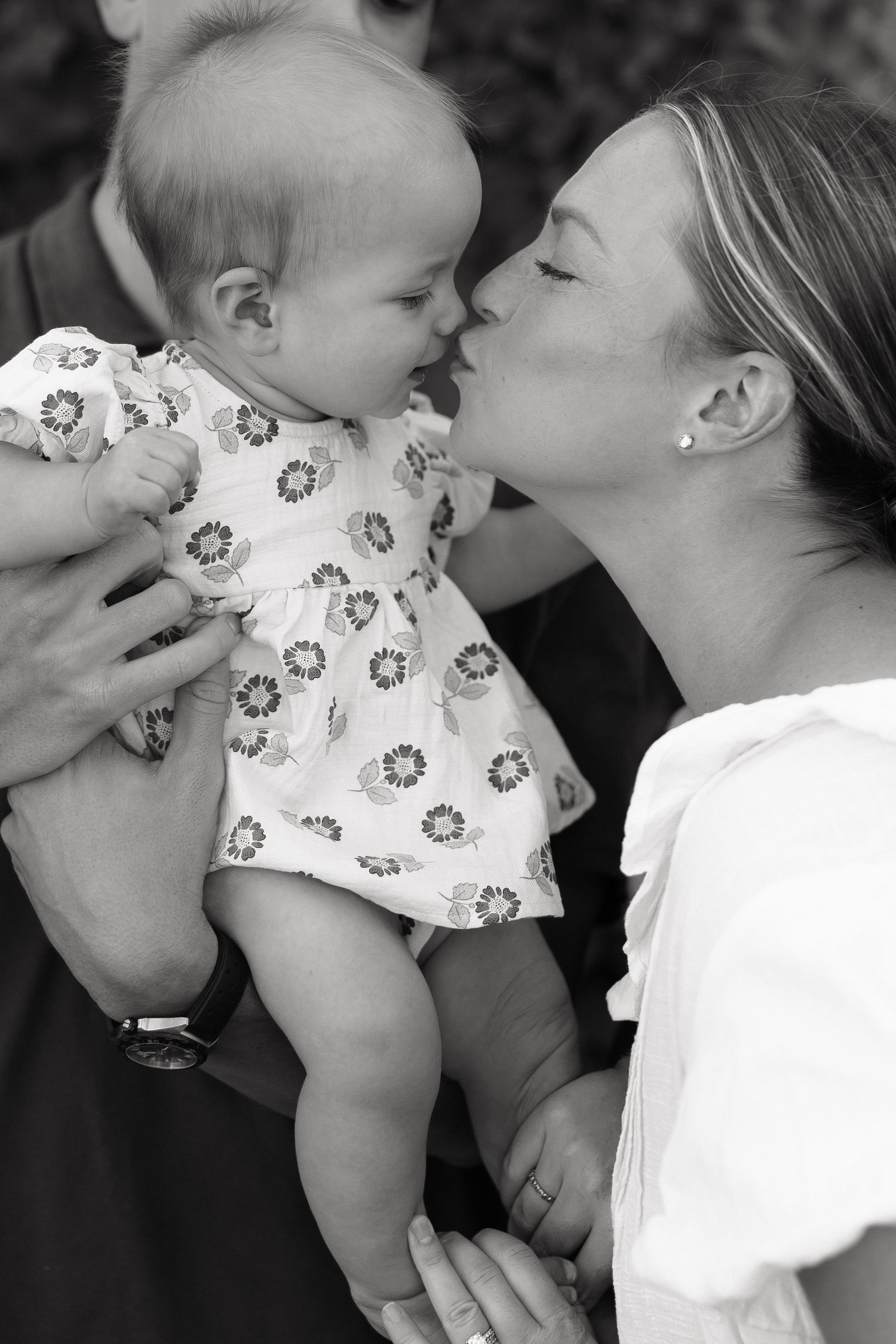 Black and white photo of a woman in white dress kissing a smiling baby in floral outfit held by another person.