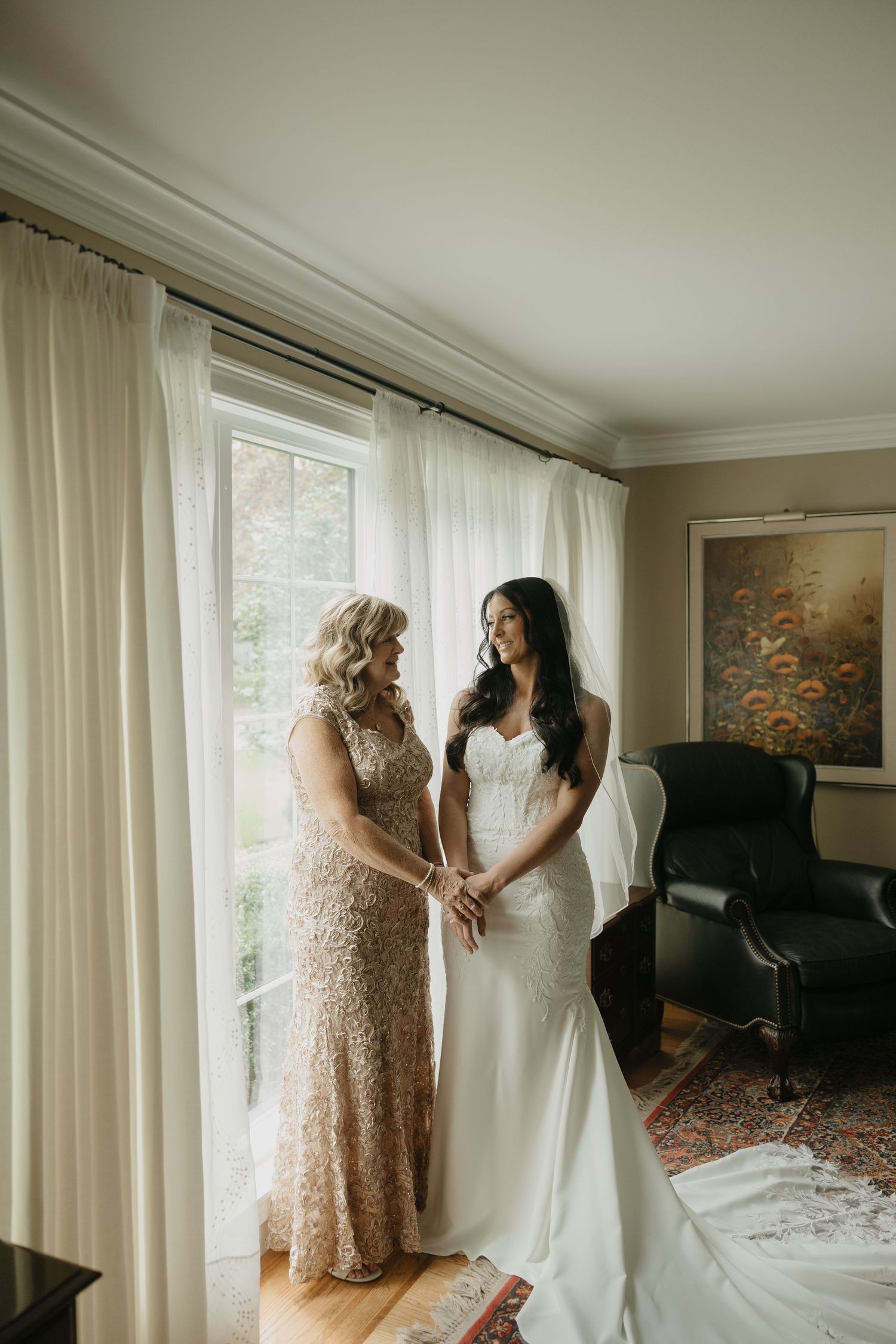 Bride in white gown stands with mother in gold lace dress by a sunlit window in an elegant room.