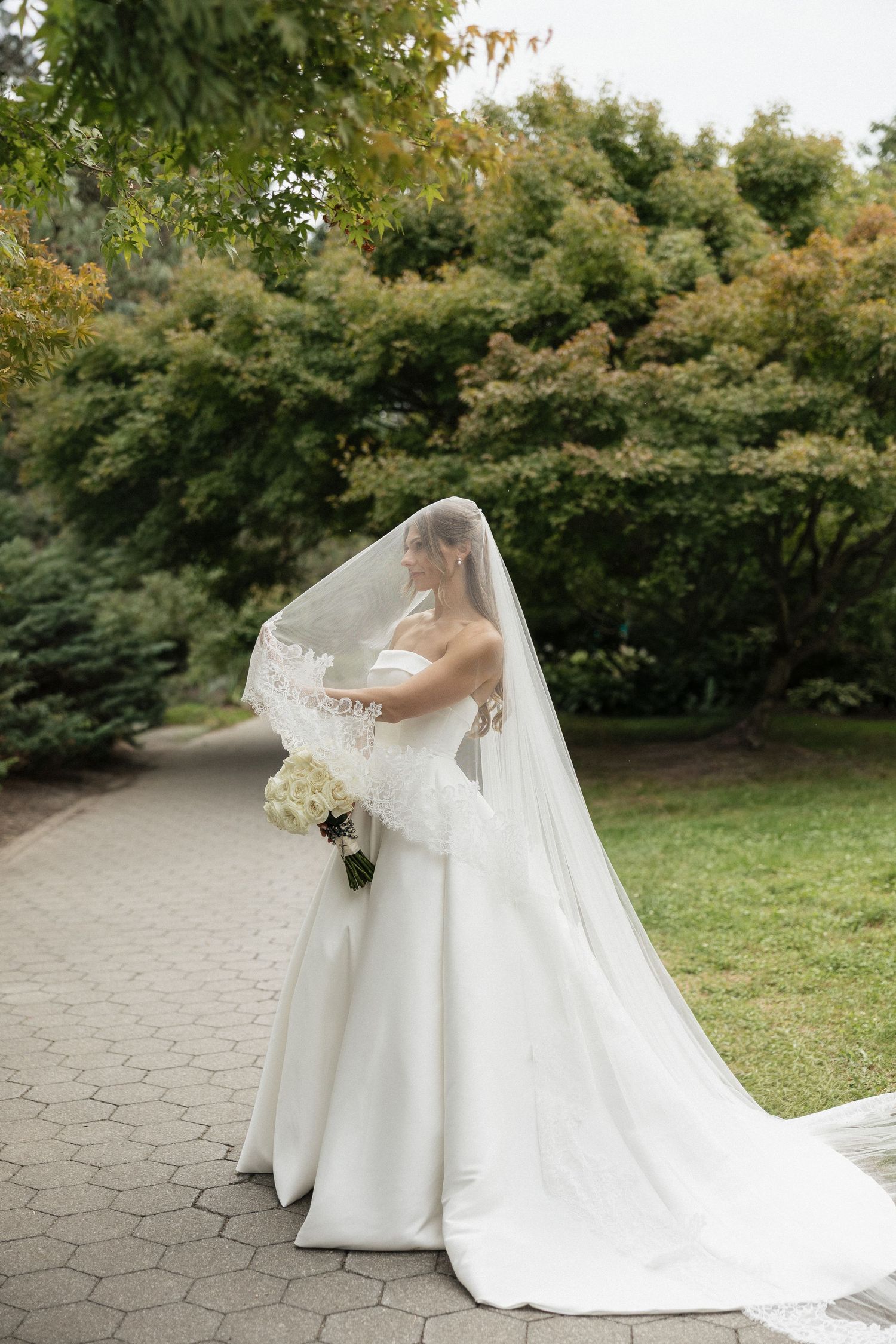 Bride in a white strapless gown with flowing veil and bouquet standing on a garden path surrounded by lush green trees.