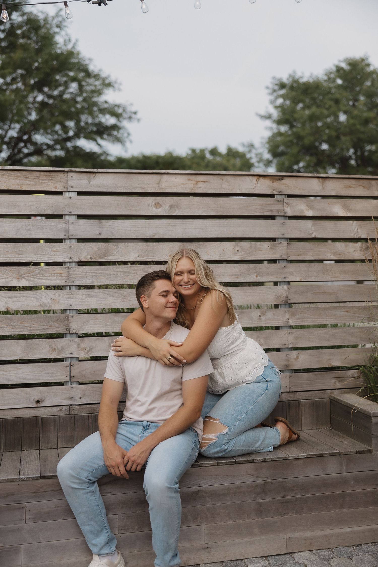 A smiling couple sitting on outdoor steps, woman hugging man from behind, wooden fence and trees in background.