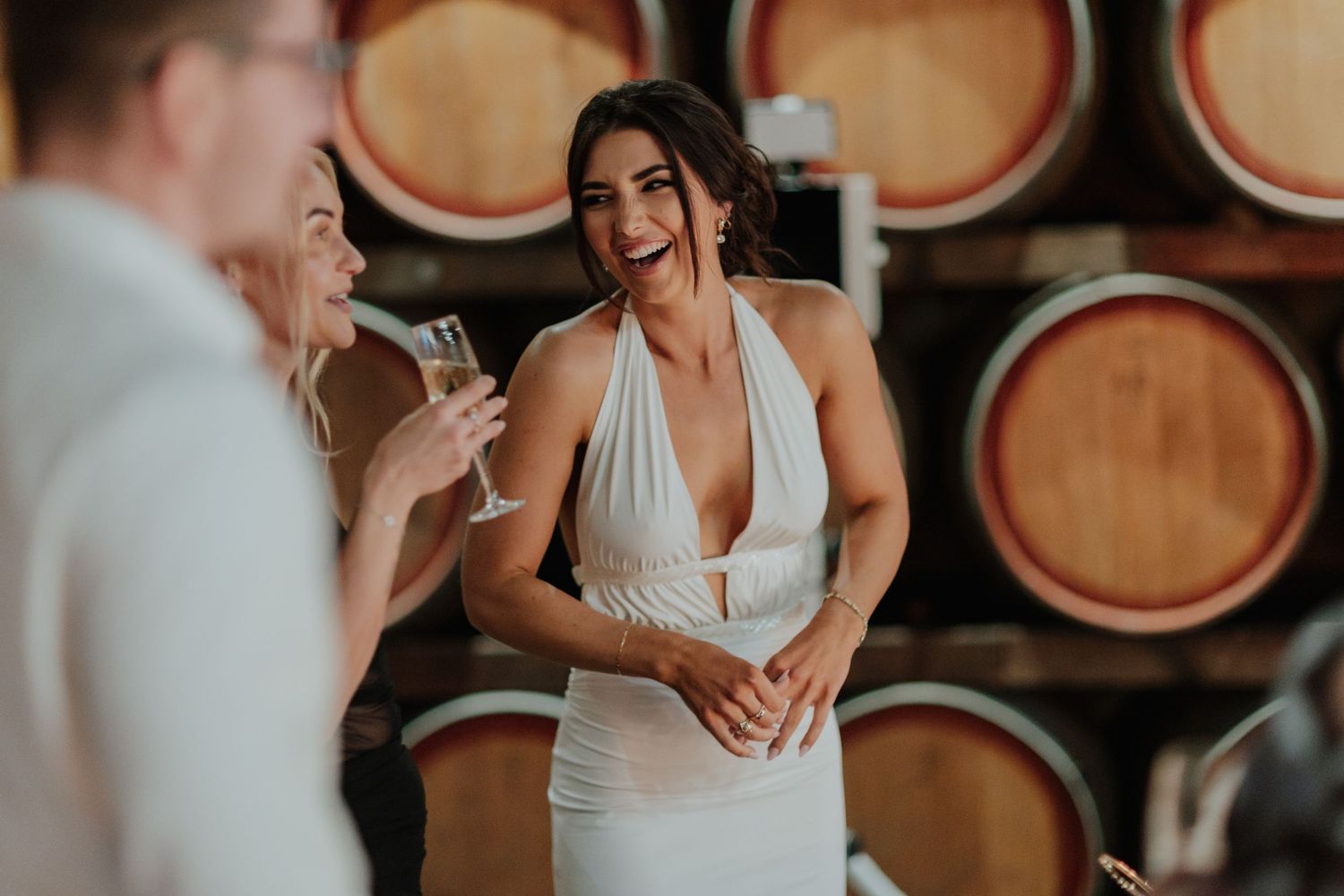 Woman in white halter dress laughing joyfully at a winery barrel room celebration.
