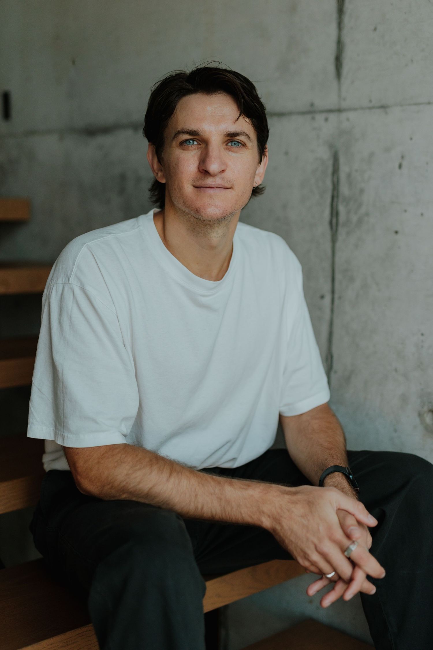 Young man in white t-shirt sitting on wooden stairs against concrete wall, smiling.
