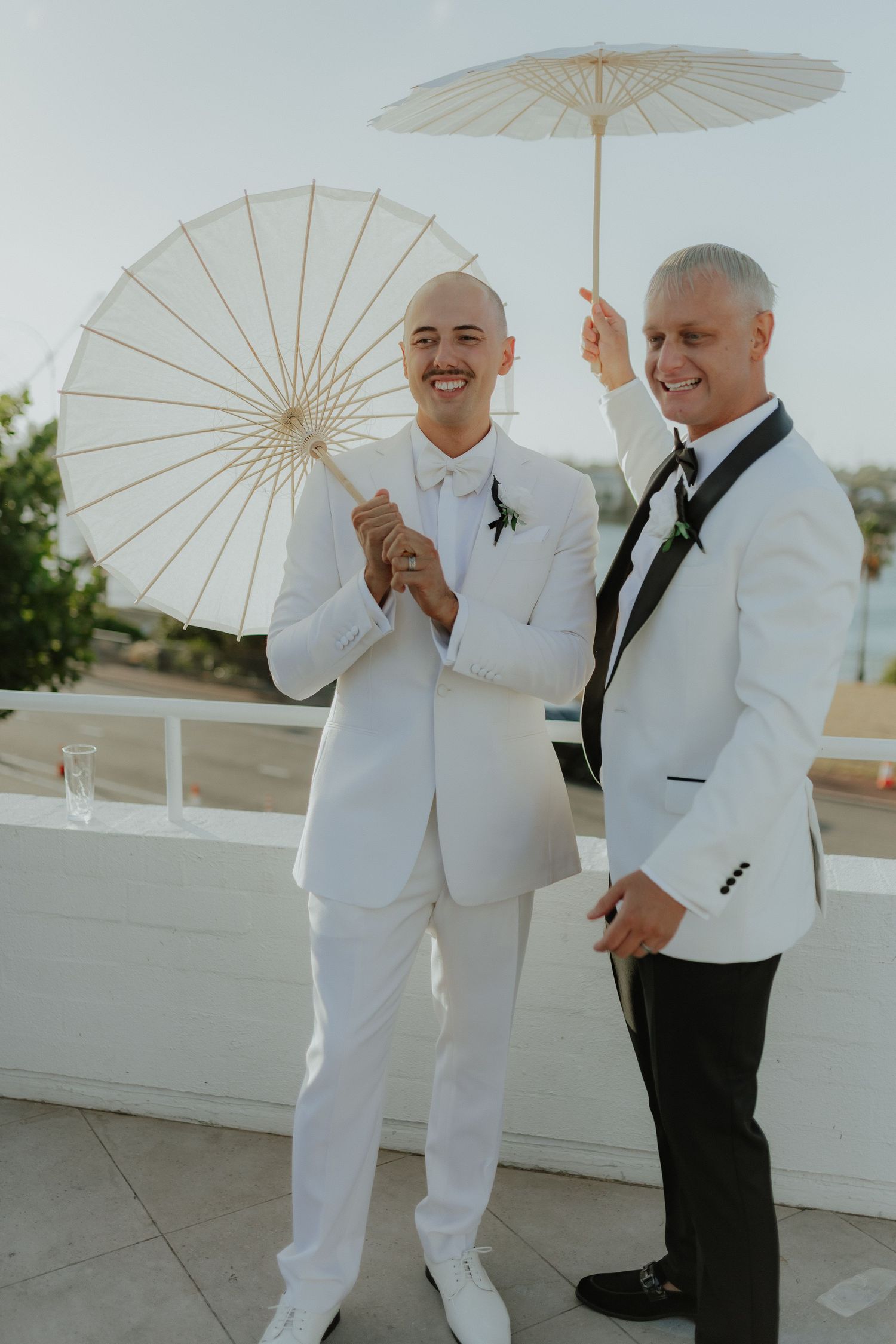Two grooms in white tuxedos smiling and holding white parasols at an outdoor wedding venue.