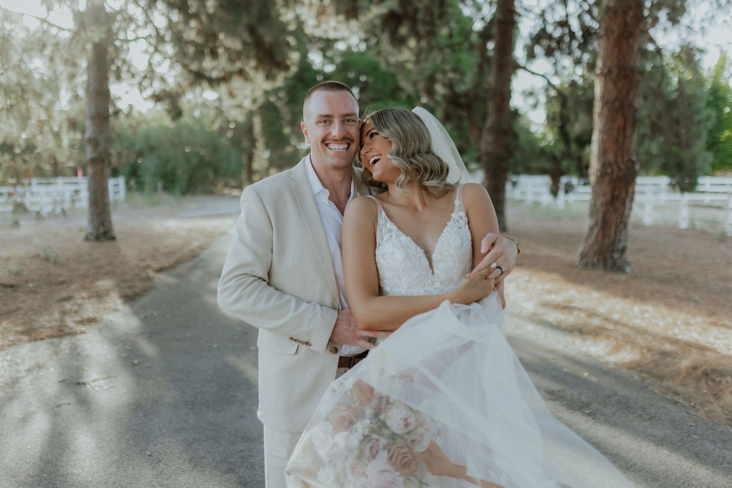 Groom in beige suit and bride in lace gown embracing warmly on a tree-lined wedding venue path.