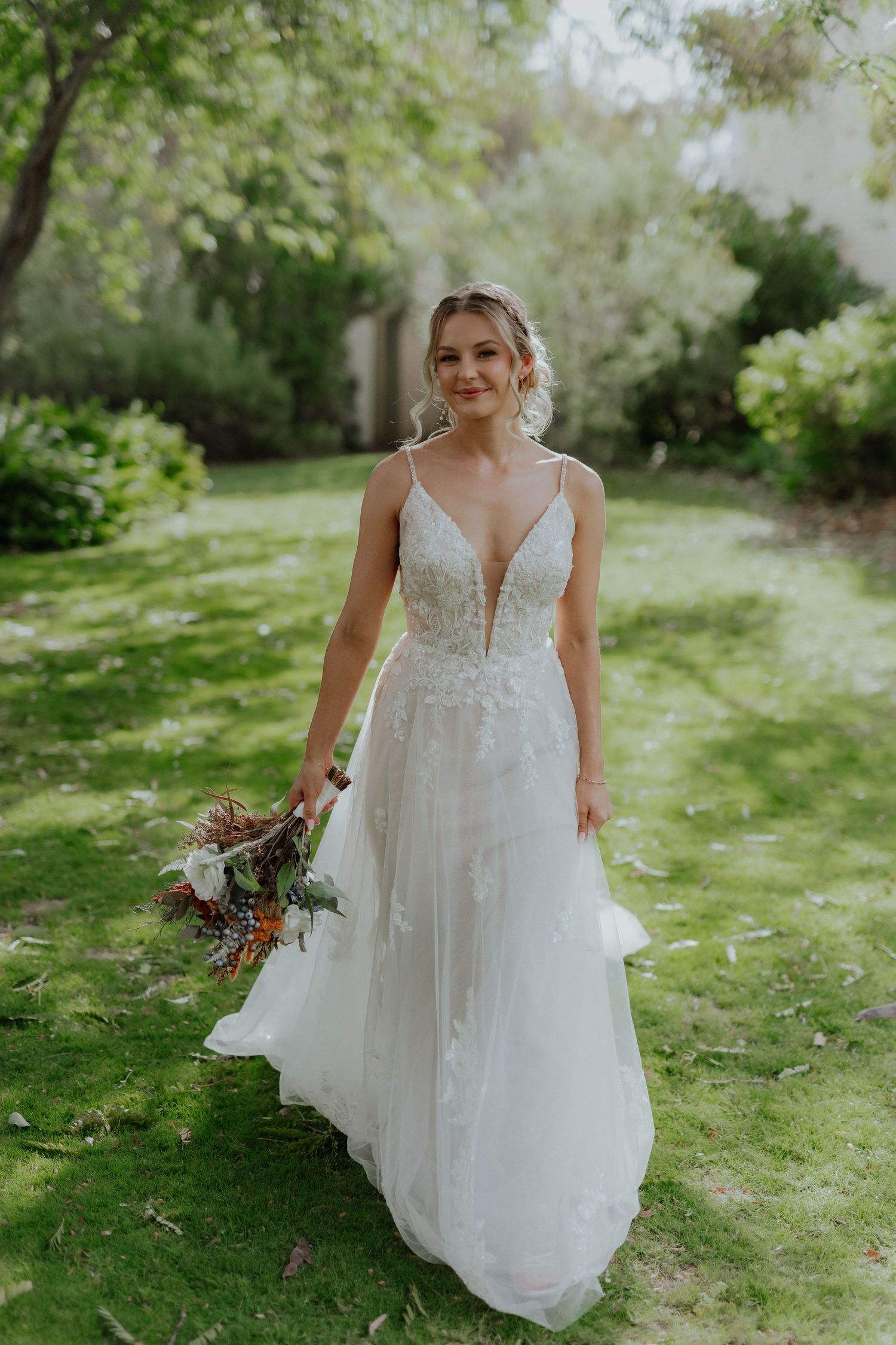 Smiling bride in floral lace A-line gown holds colorful bouquet in lush garden setting.