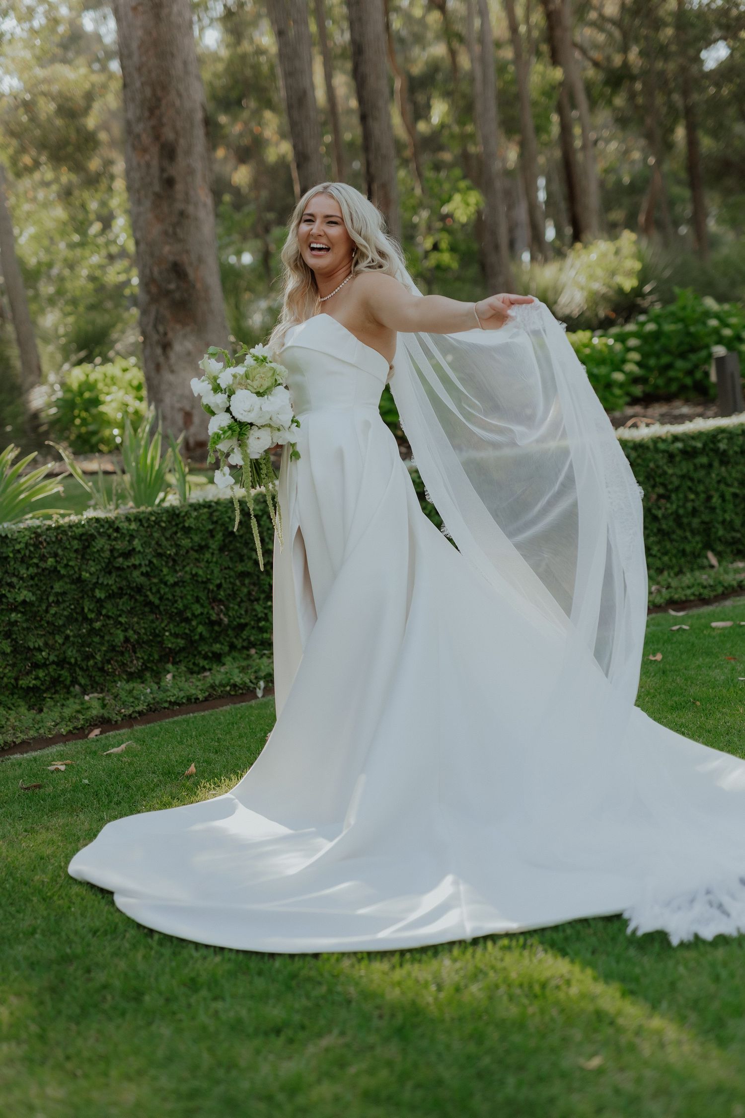 Joyful bride in white strapless gown twirls veil holding bouquet in sunlit garden setting.