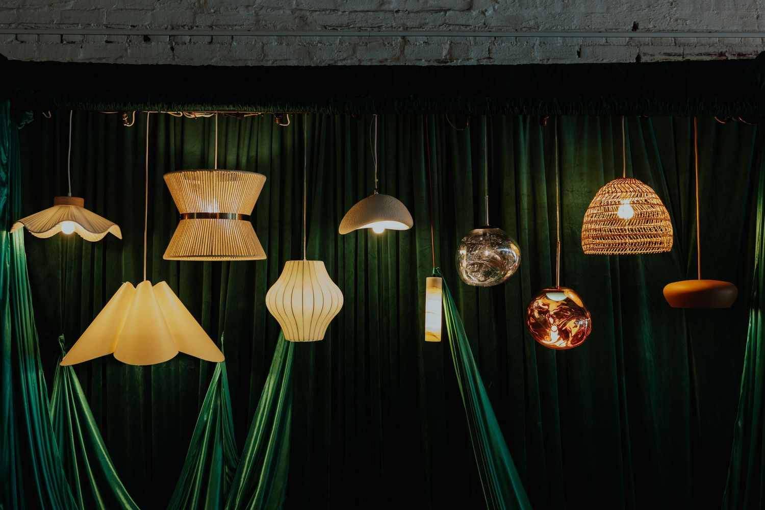 Collection of diverse hanging pendant lamps against lush green velvet curtain backdrop.