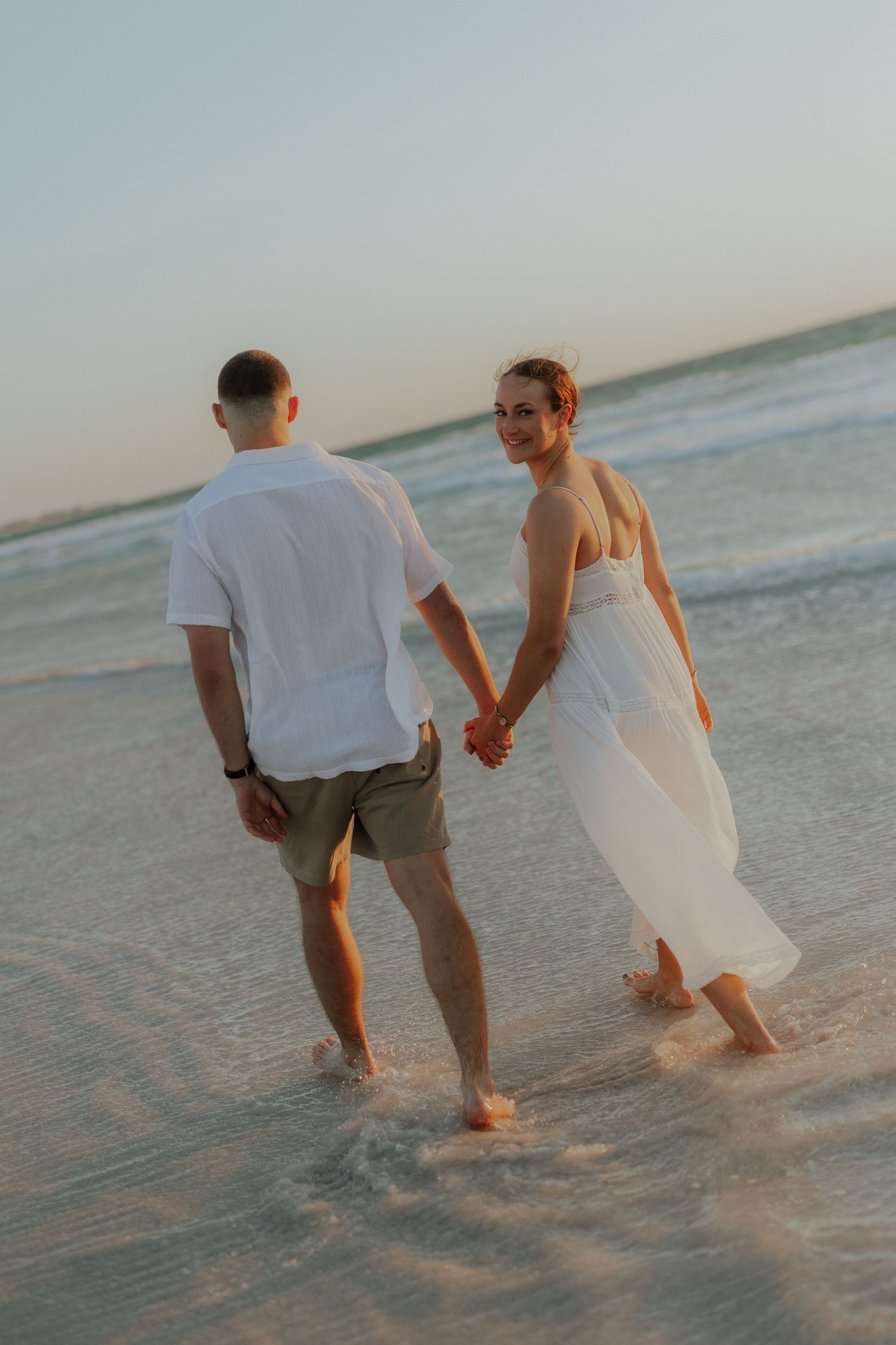 Couple holding hands wading in ocean waves at sunset, woman in white dress looking back smiling.
