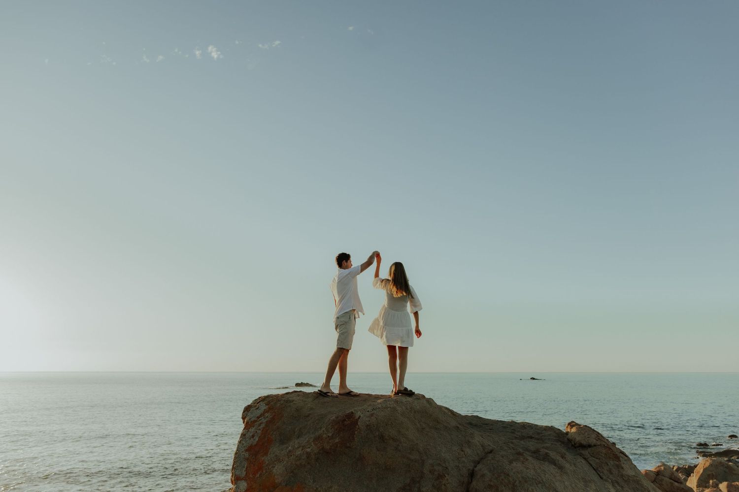 Romantic couple dancing on a large rock with the ocean horizon and clear sky behind them.
