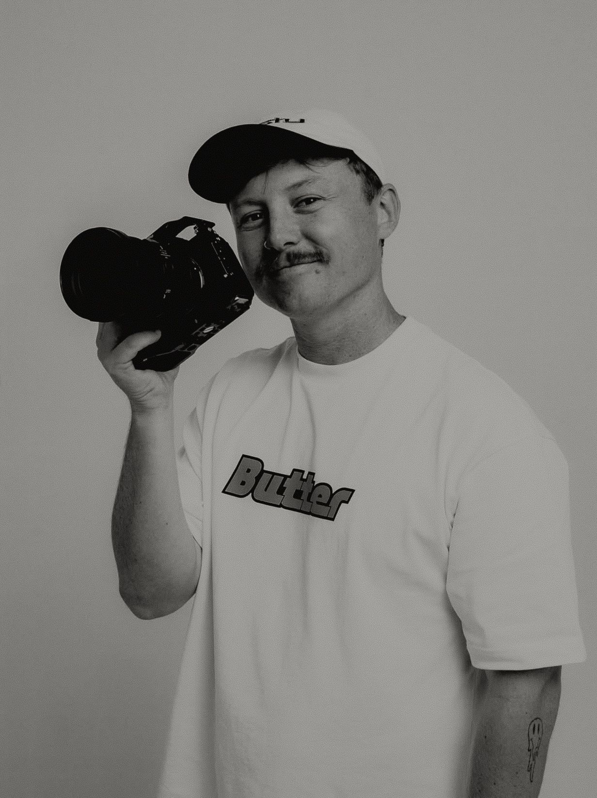 Man in white Butler t-shirt and cap holding professional camera in black and white portrait.