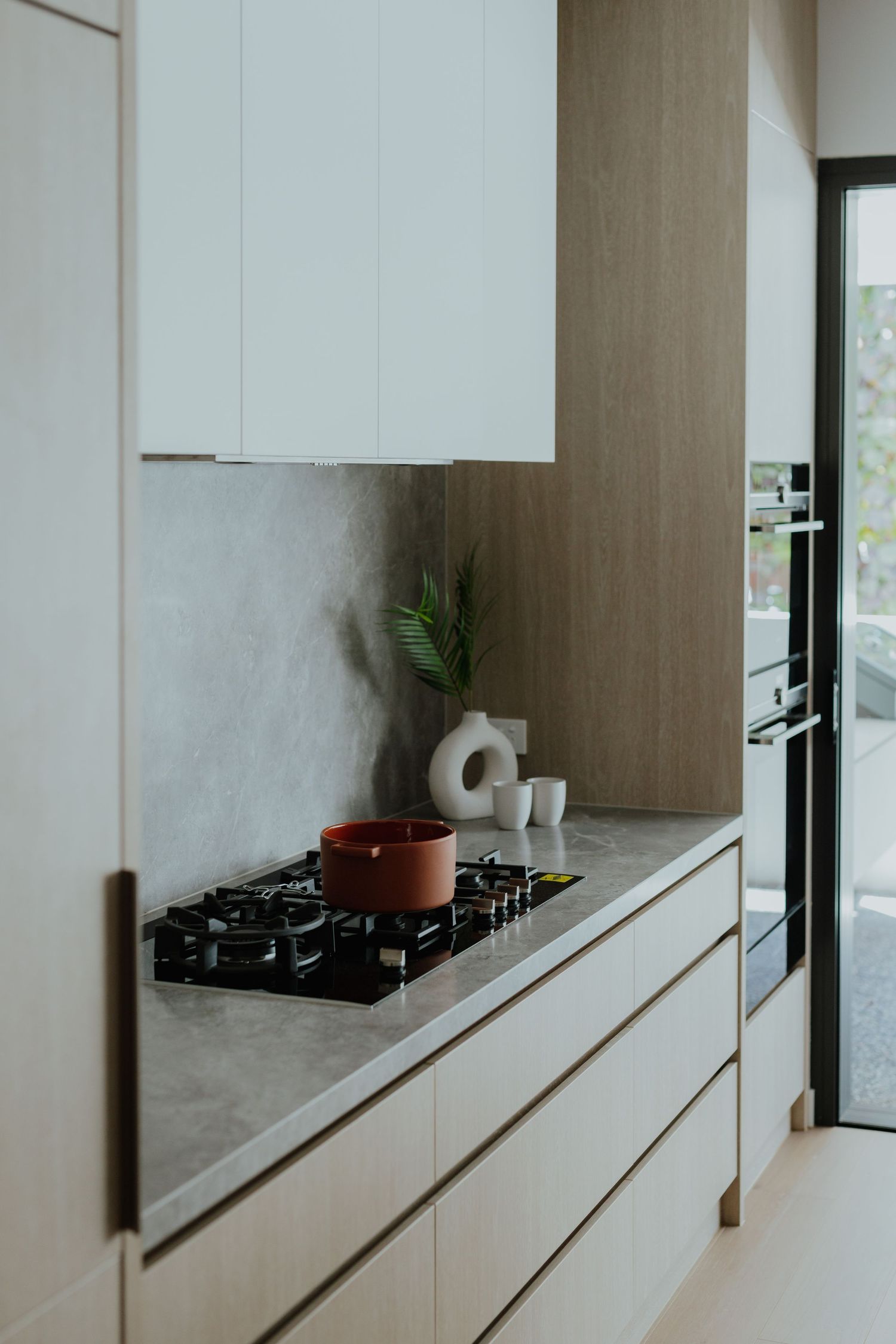 Modern white kitchen with gas cooktop, grey stone countertops, handleless cabinets, and decorative vase with plant.