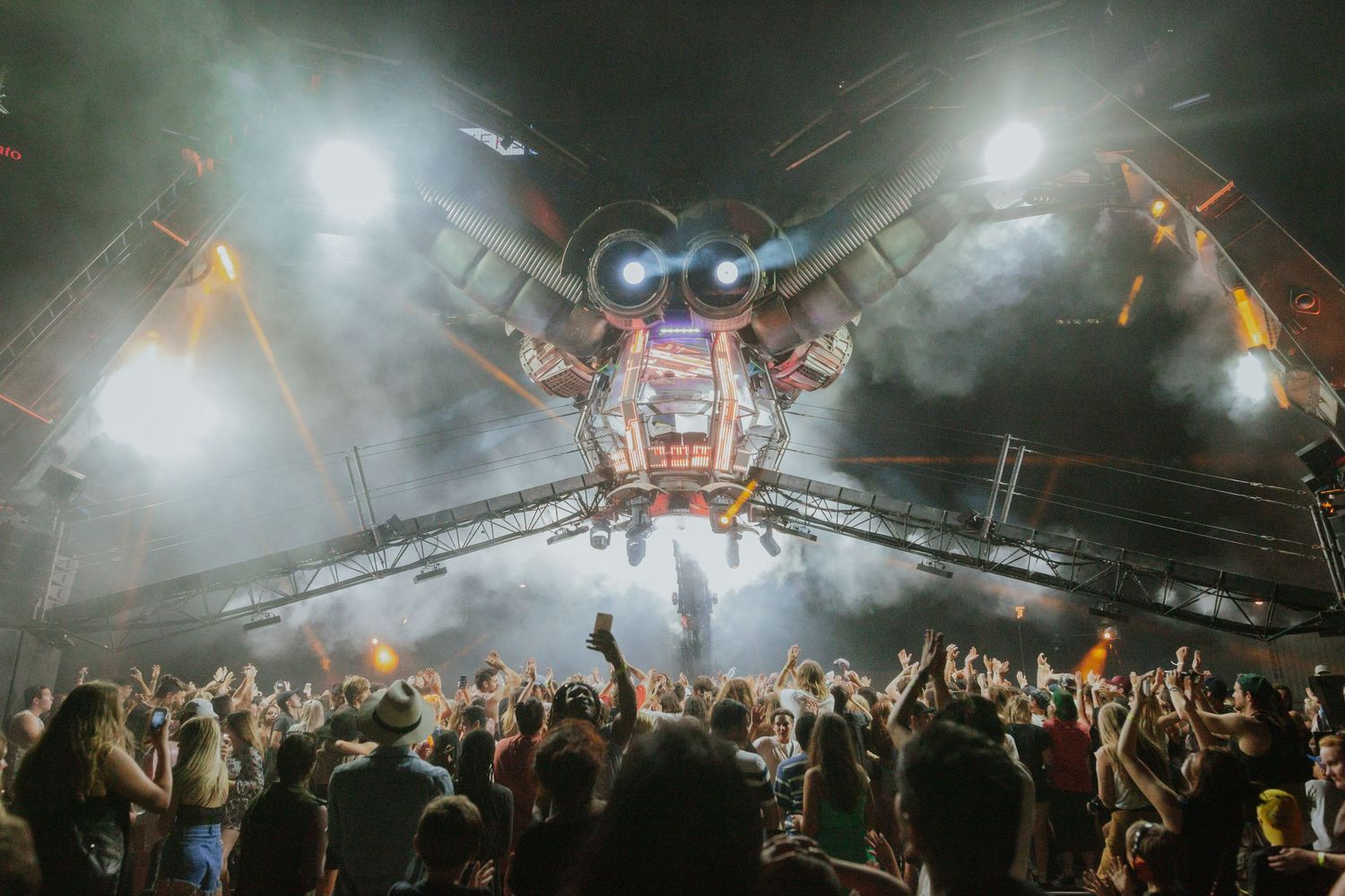 Massive crowd cheers beneath a dramatic mechanical stage structure with bright lights at a music festival.