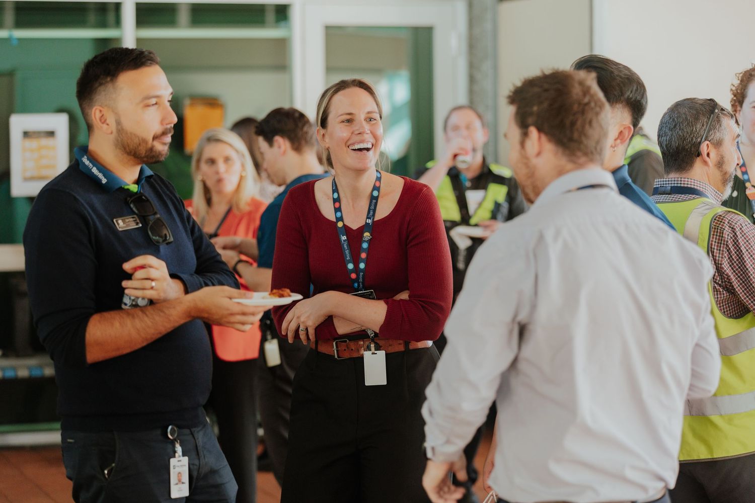 Colleagues networking and laughing at a corporate workplace event wearing lanyards and hi-vis vests.