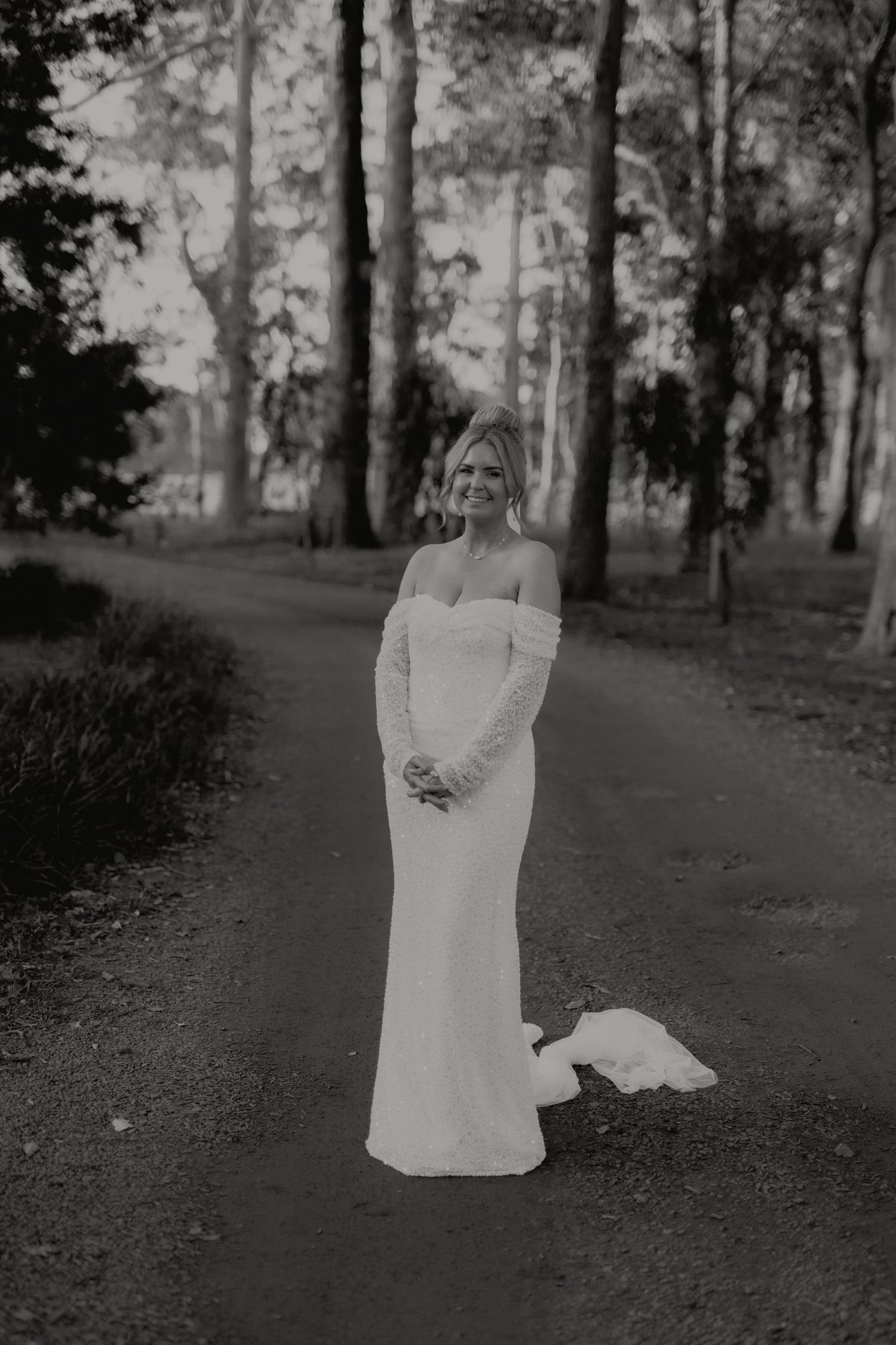 Black and white portrait of bride in off-shoulder lace gown standing on forest pathway.
