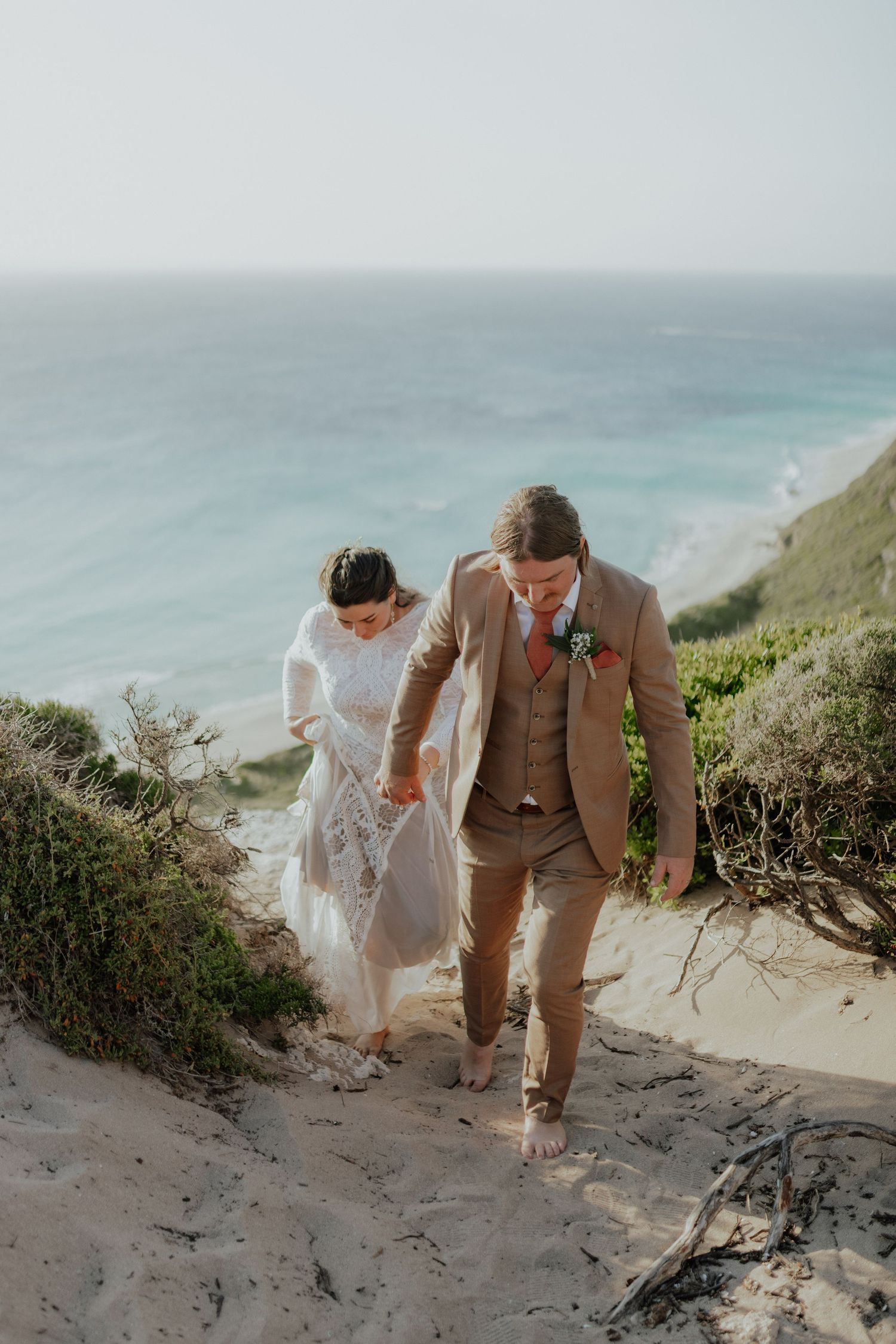 Barefoot bride and groom walk down sandy coastal dunes with turquoise ocean backdrop.