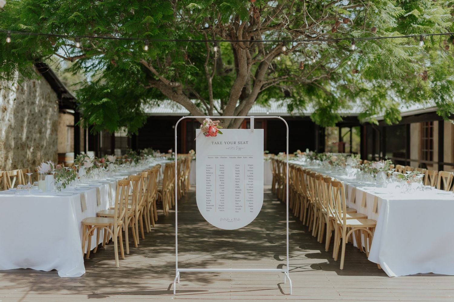 Outdoor wedding reception with long white tables, wooden chairs, and a floral seating chart display under a tree.