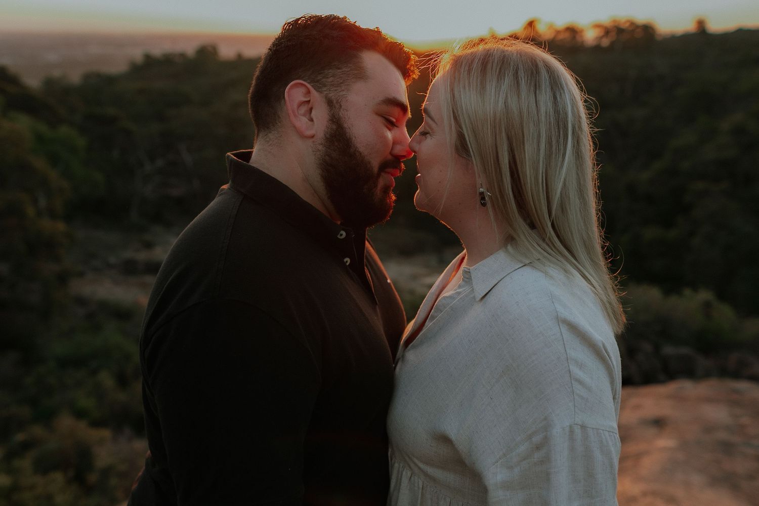 Romantic couple almost kissing at golden hour with lush green hills and sunset sky in the background.