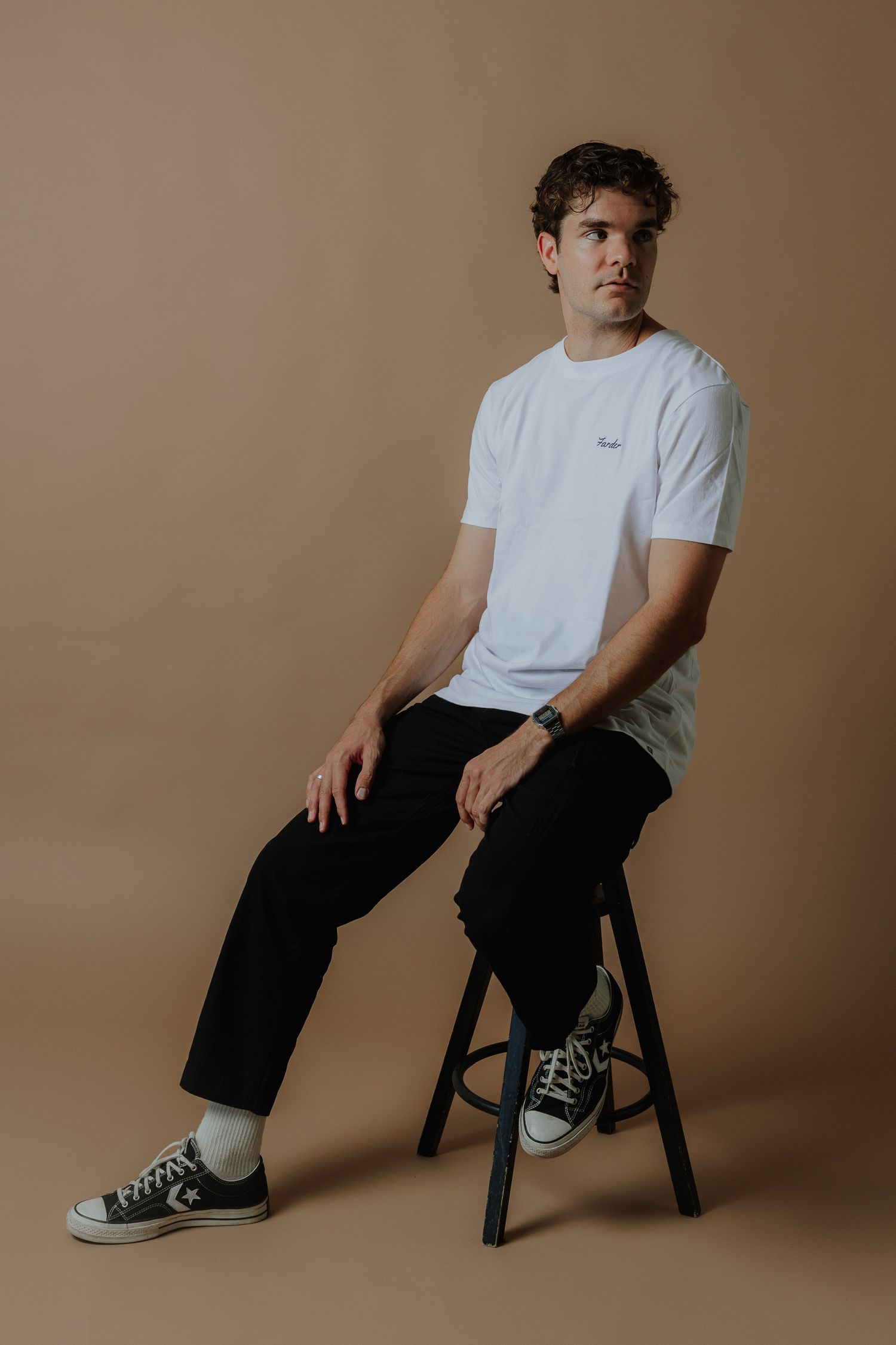 Young man in white t-shirt, black pants, and Converse sneakers sitting on stool against beige background.