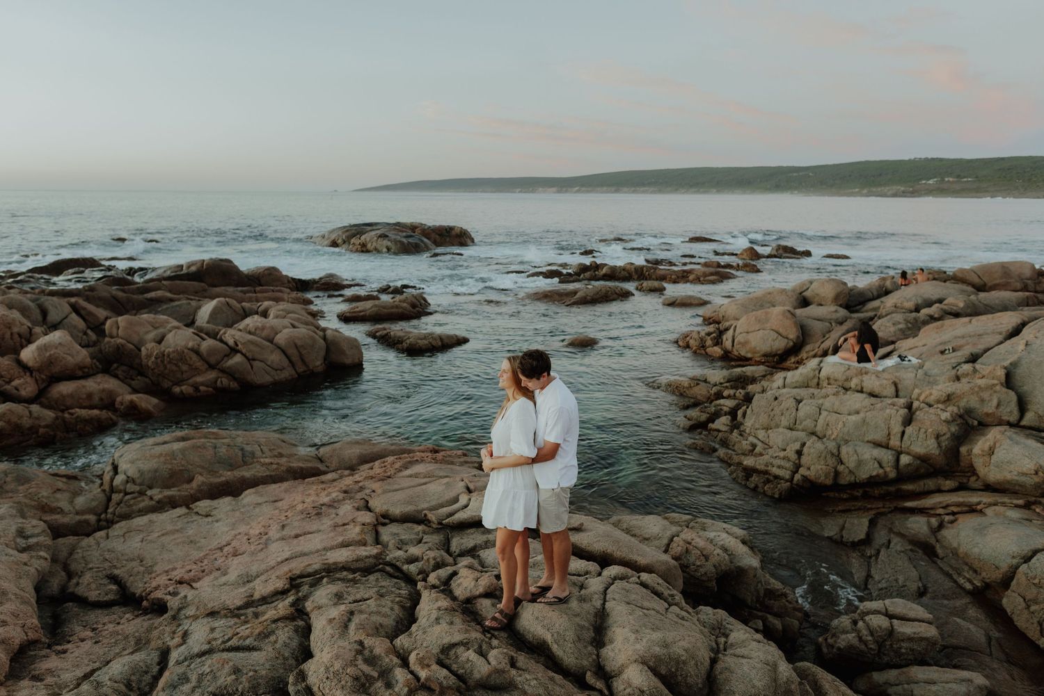 Couple embracing on rocky coastline at dusk with calm ocean and green headland in background.