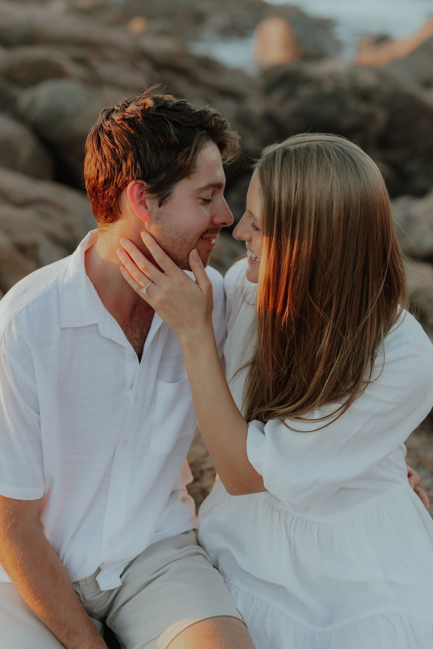 Close-up of smiling couple about to kiss during golden hour engagement shoot on rocky beach.