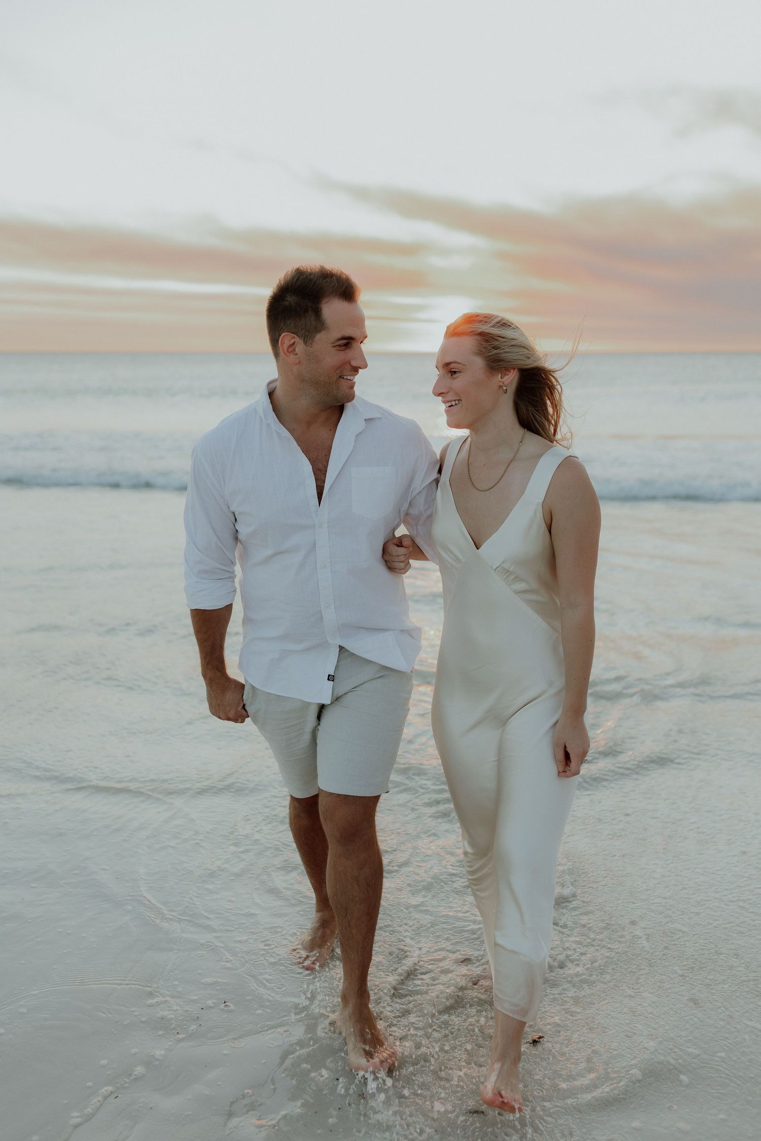 Romantic couple walking hand in hand through ocean waves at sunset, dressed in white and cream outfits.