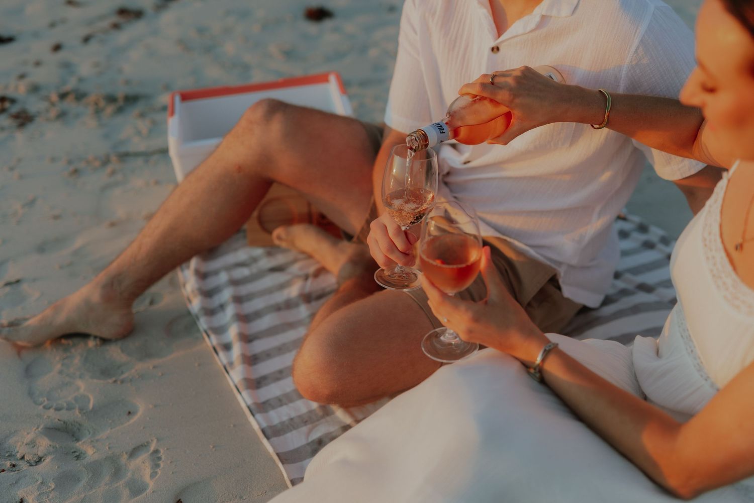 Couple clinking wine glasses on a beach blanket at golden hour.