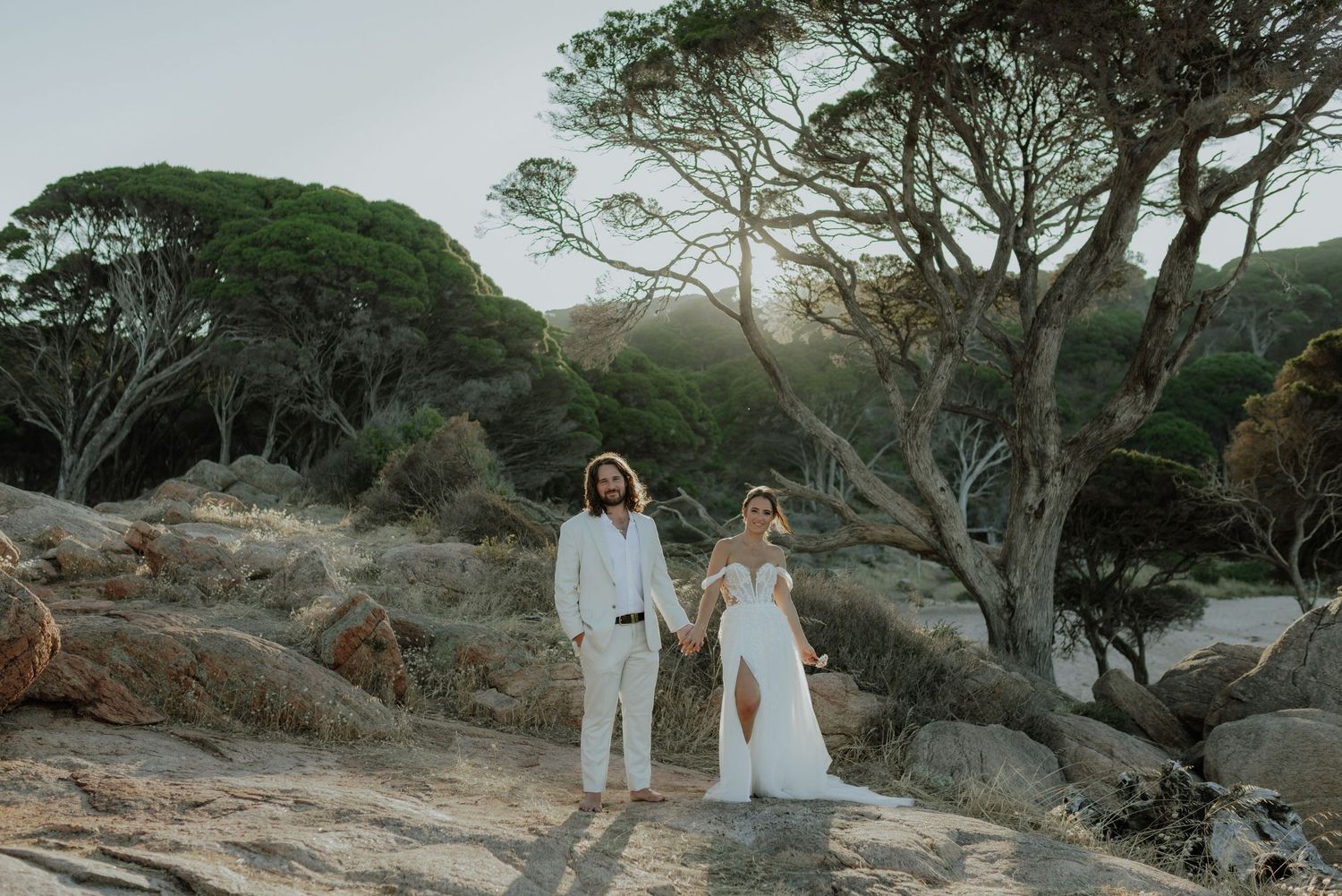 Bride and groom holding hands on rocky coastal landscape with pine trees at golden hour.