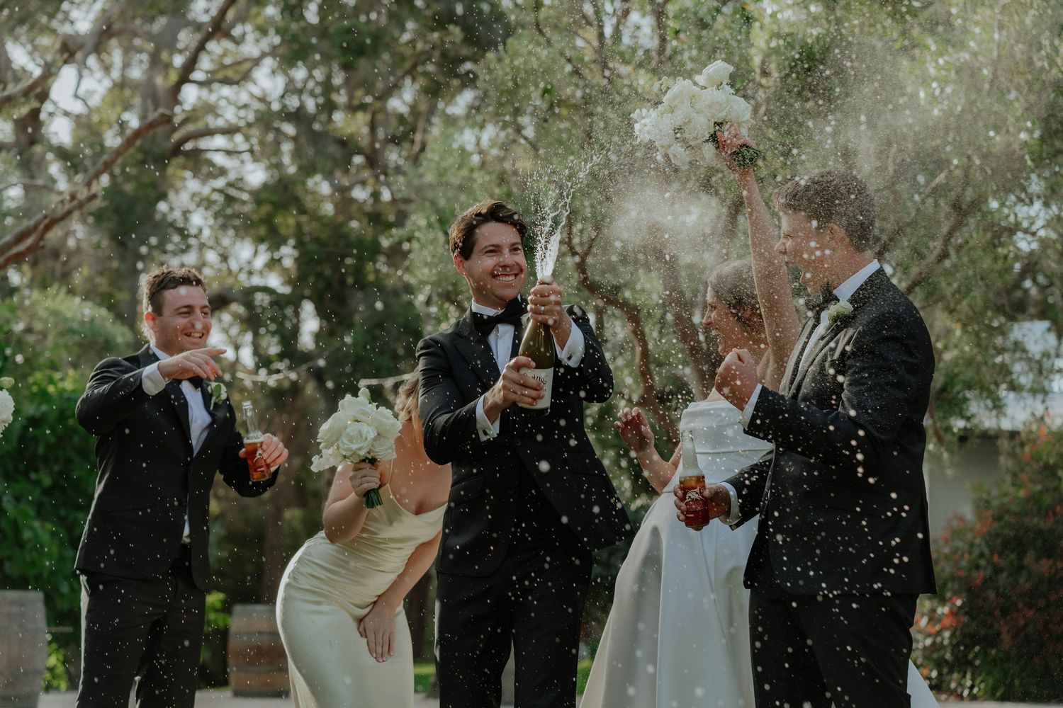 Wedding party celebrating with champagne spray outdoors, groom in black tuxedo surrounded by guests and bride.