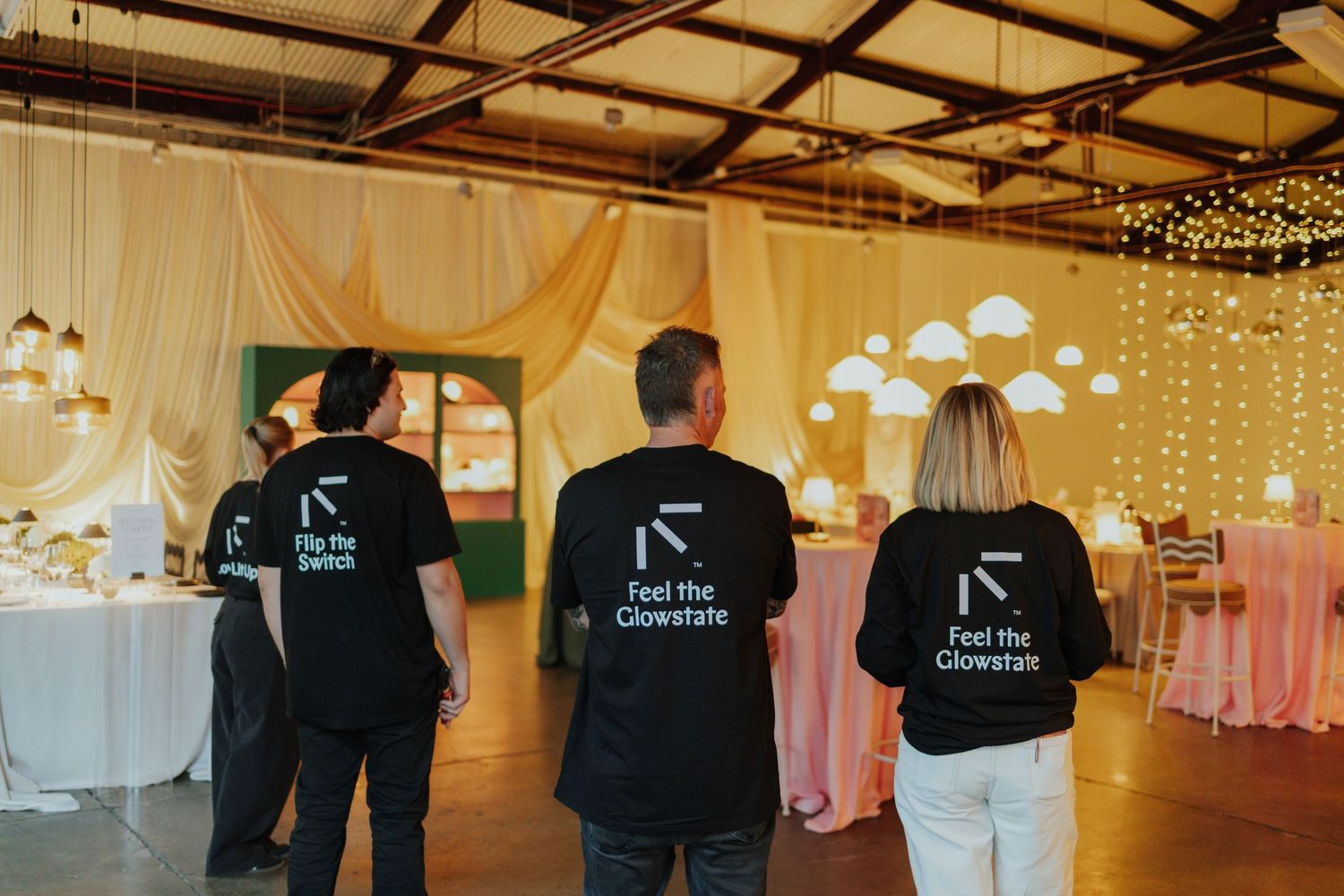Three event staff in black Feel the Glowstate branded jackets standing in a decorated wedding venue.