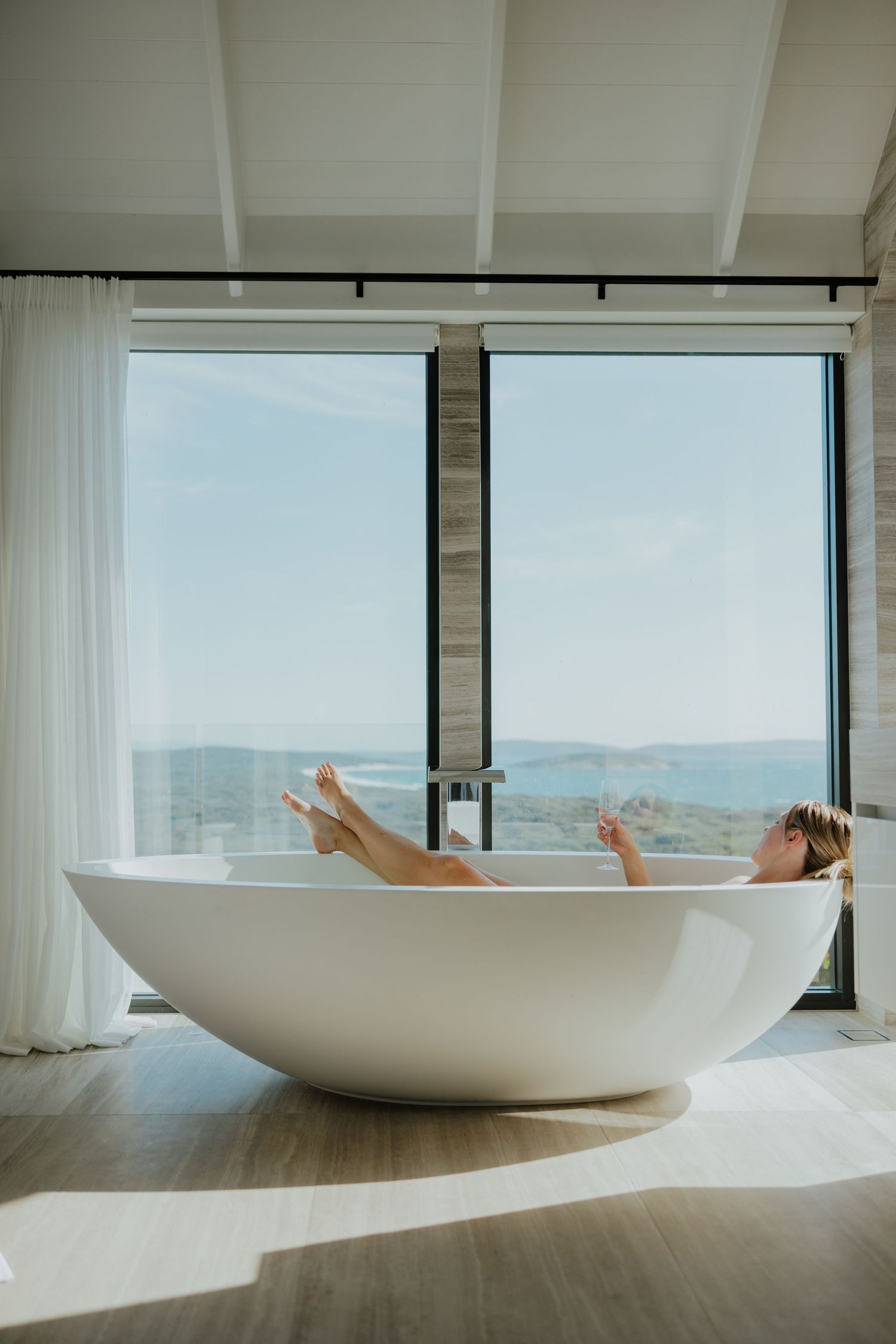Woman relaxing in freestanding white bathtub with panoramic ocean view window.