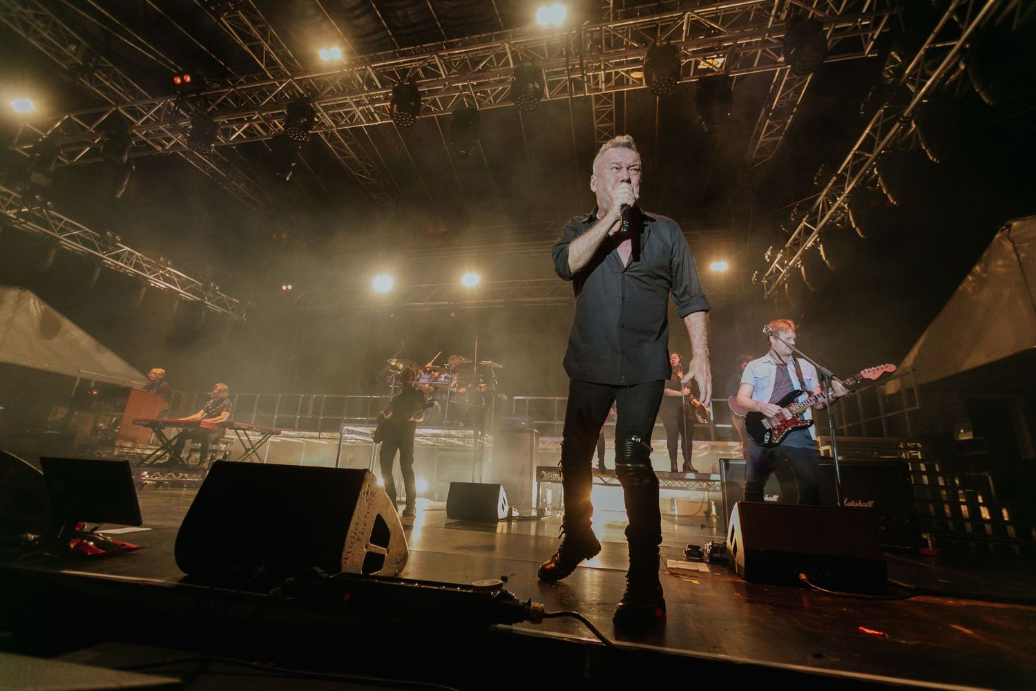 Male vocalist singing on stage with band members and dramatic stage lighting at live concert.