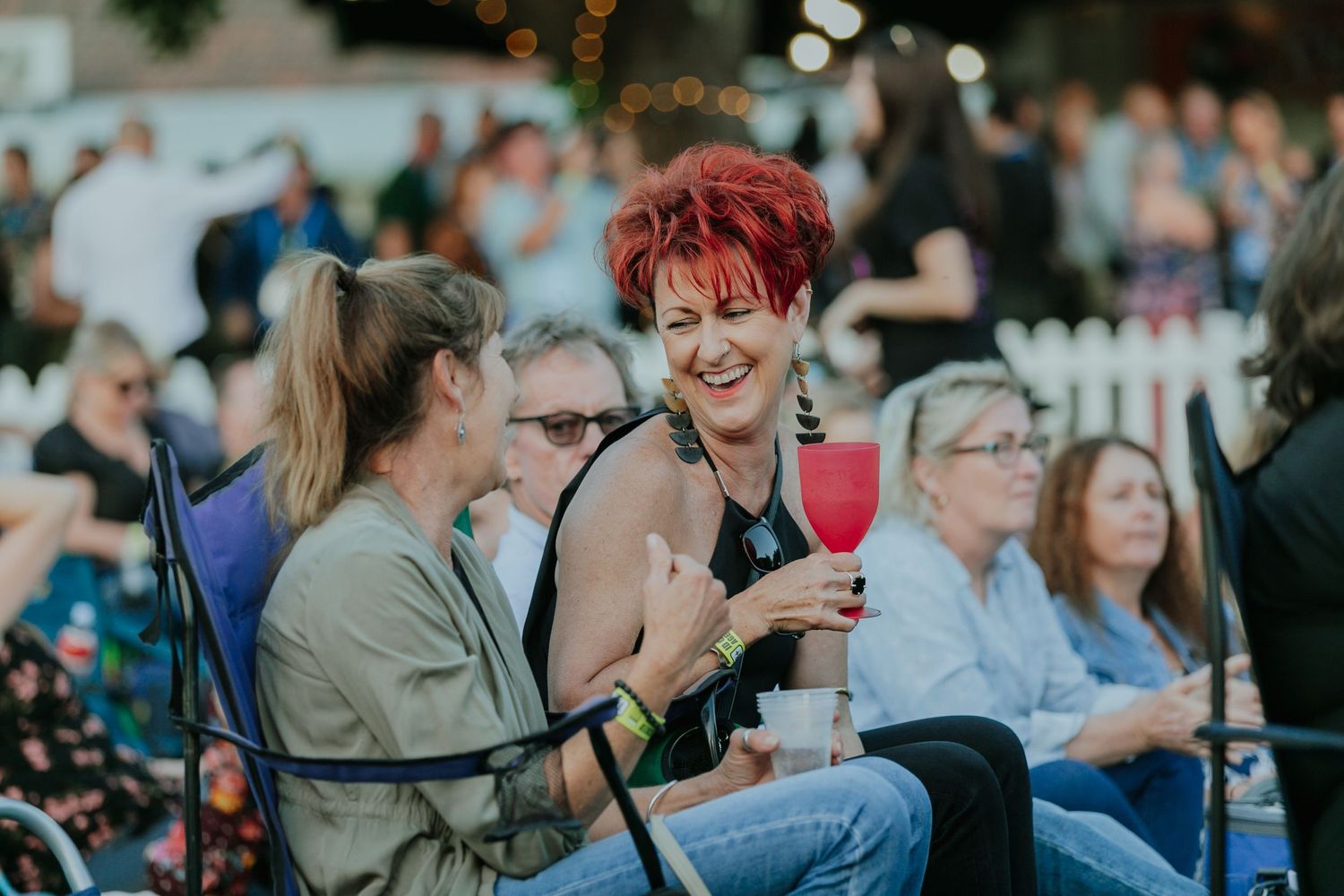 Two women laughing and socializing at an outdoor music festival, one holding a red drink.