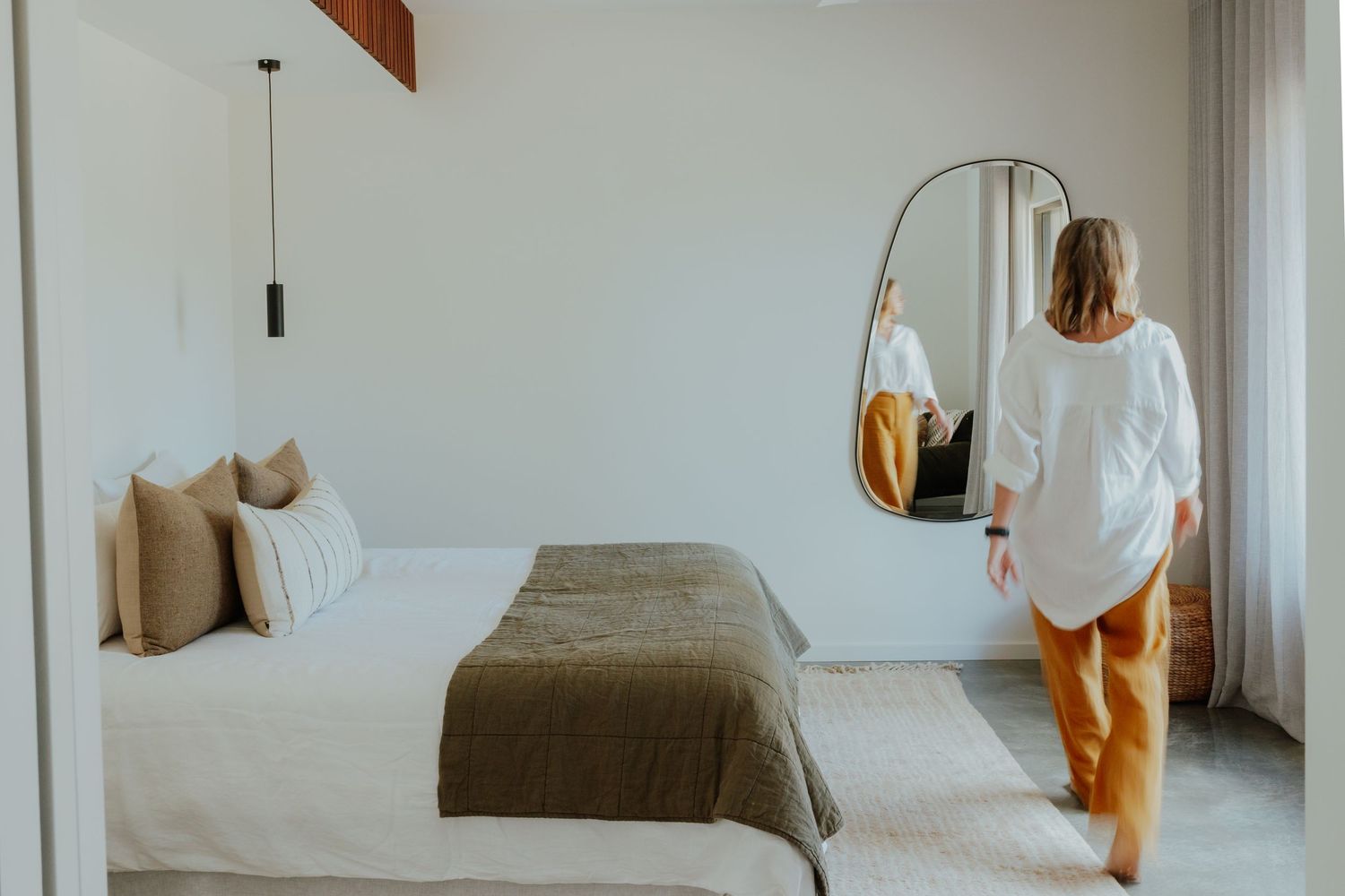 Woman standing in a bright minimalist bedroom with white linen, olive throw and an arched full-length mirror.