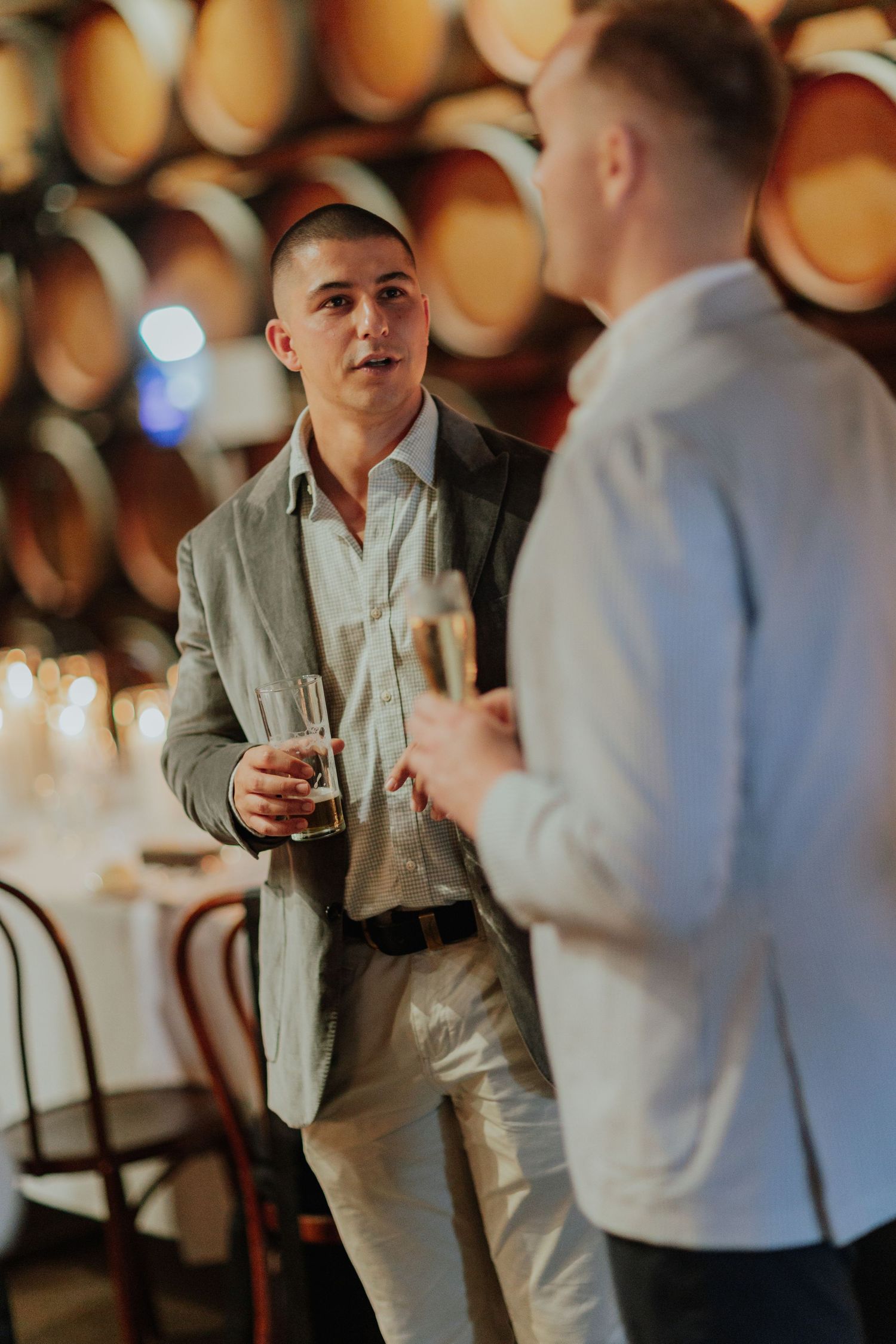 Two men in smart casual attire conversing with champagne glasses in a winery barrel room.