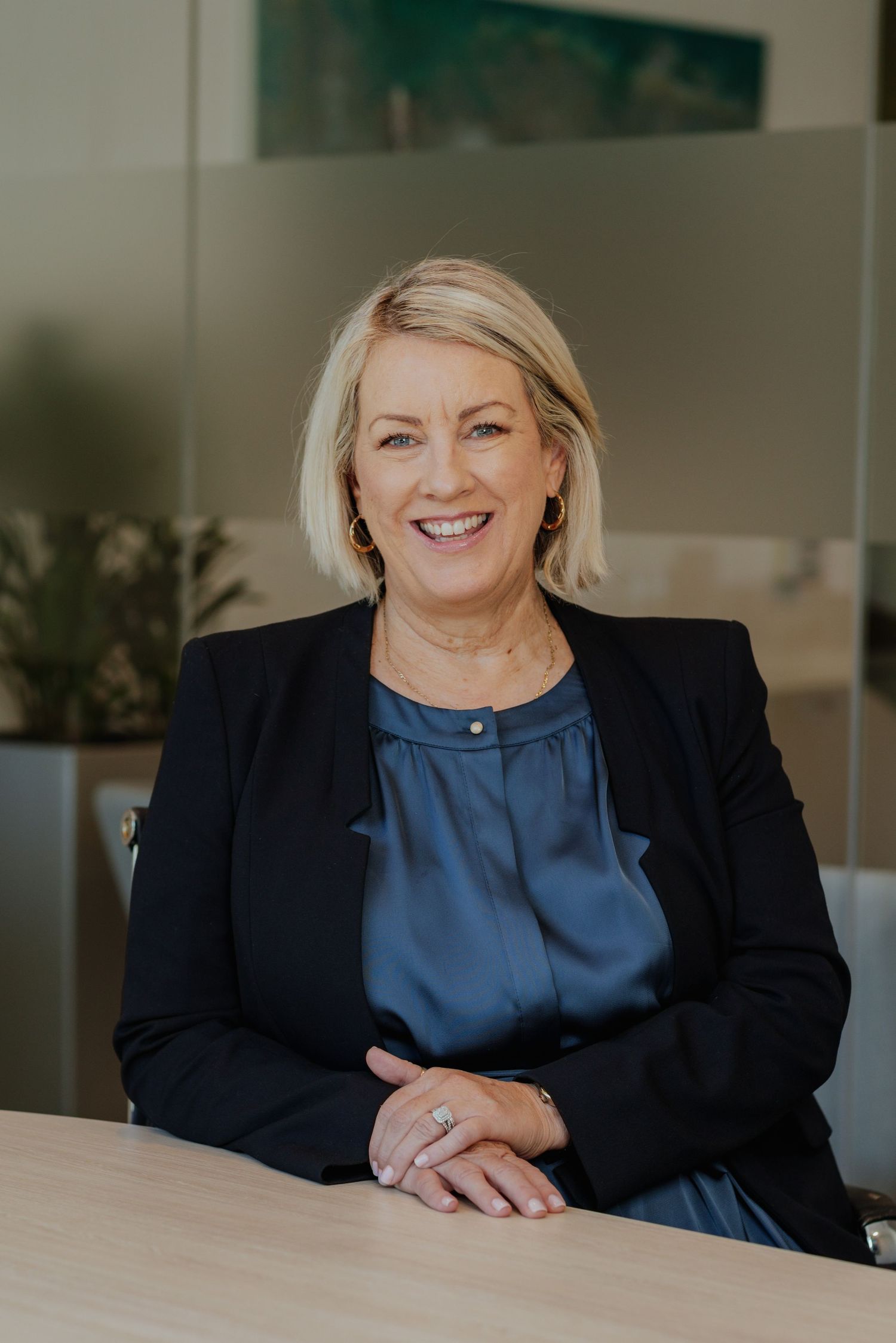 Smiling professional blonde woman in black blazer and blue top at office desk.