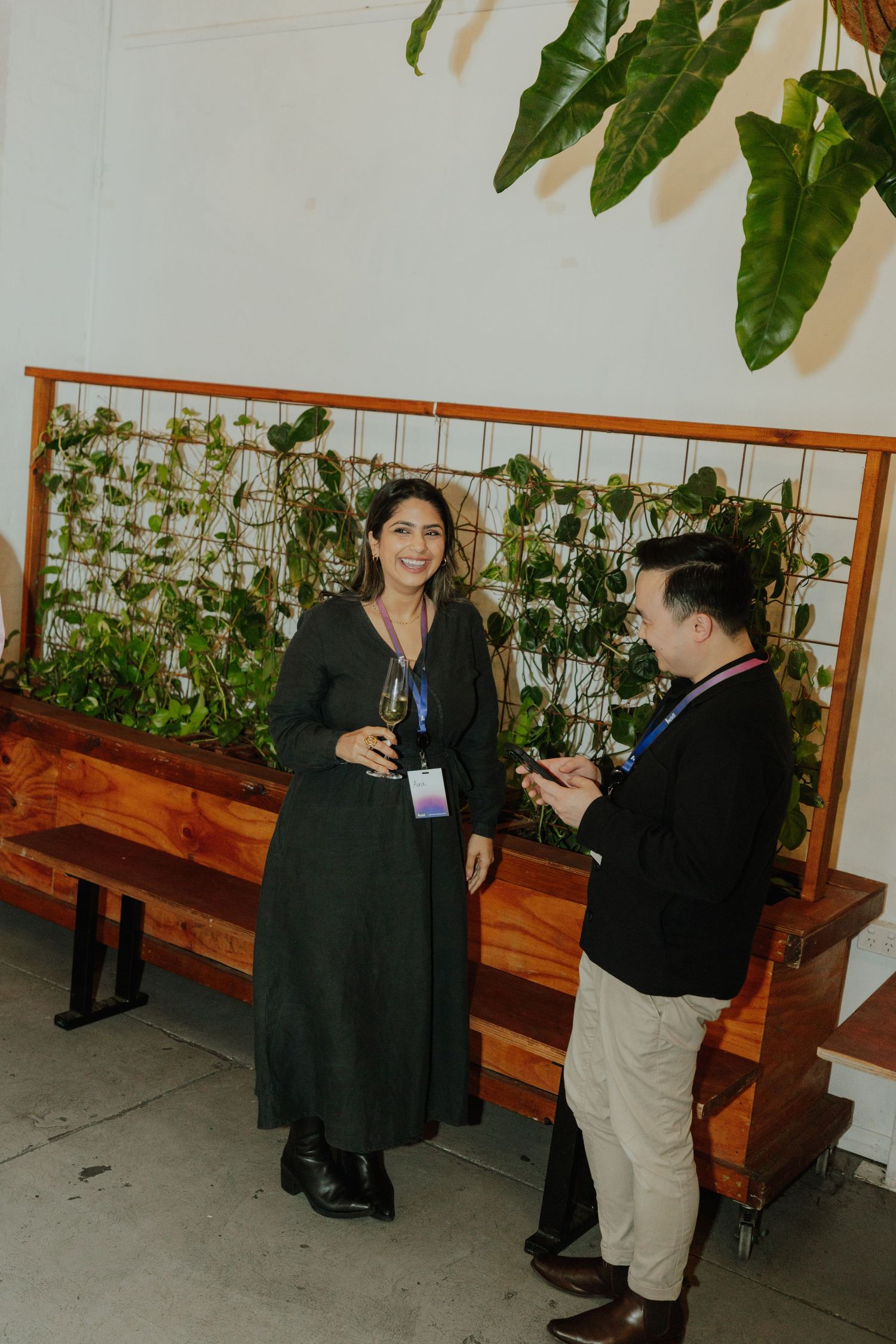 Two conference attendees with lanyards chatting near a green plant wall at an indoor event.