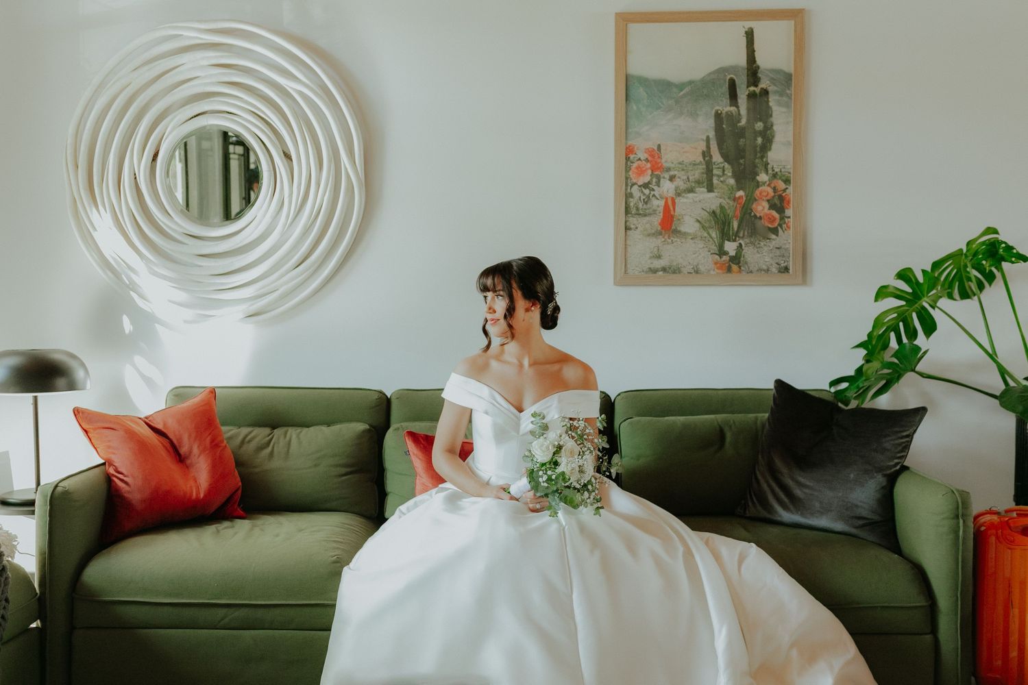 Bride in off-shoulder ball gown sits on green sofa holding bouquet, with circular mirror and cactus art on wall.