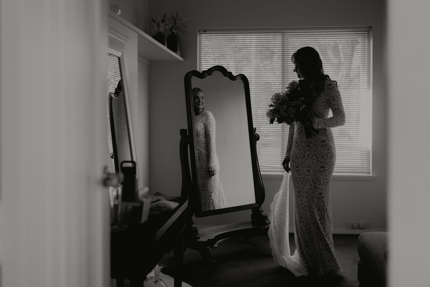 Black and white photo of a bride in a lace gown holding flowers beside a full-length mirror.