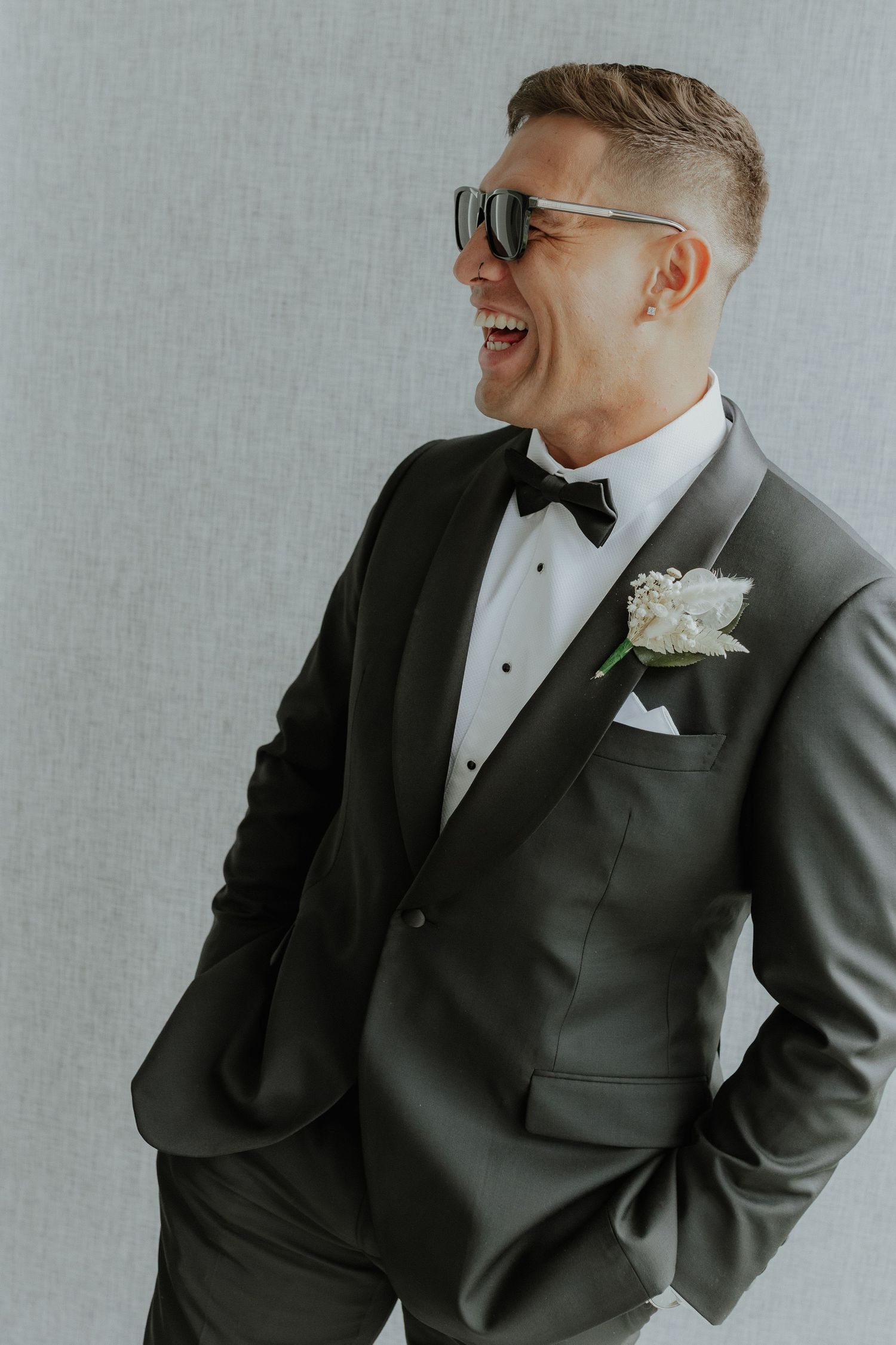 Laughing groom in charcoal tuxedo with white boutonniere and sunglasses posing against a light gray background.