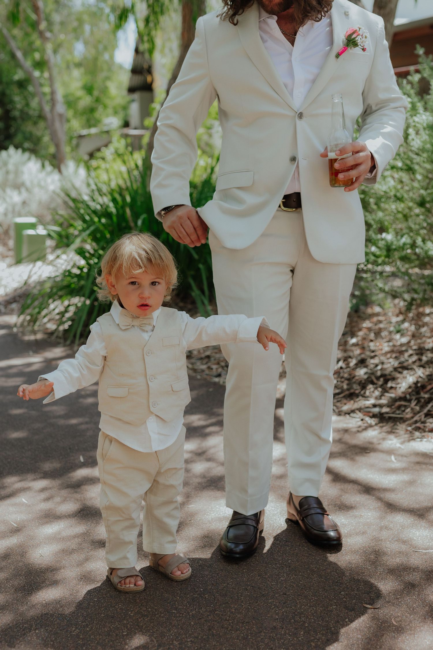 Toddler in cream suit standing beside groom in matching ivory suit at outdoor wedding.