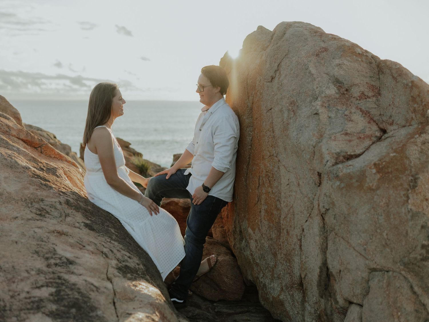 Couple sitting in rocky crevice by the ocean with warm backlit sunlight glowing behind them.