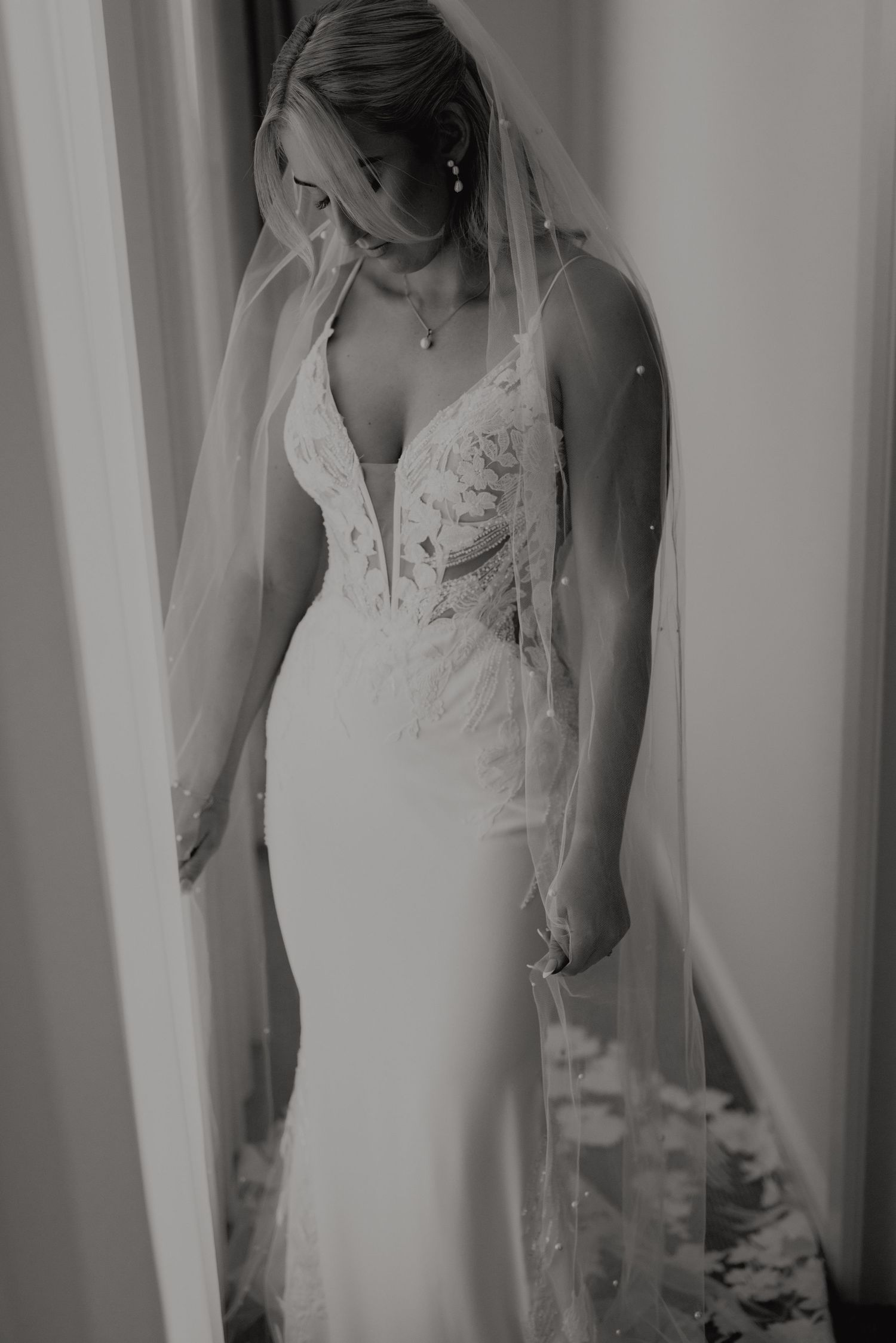 Black and white portrait of bride in deep V lace gown with flowing veil standing by a window.