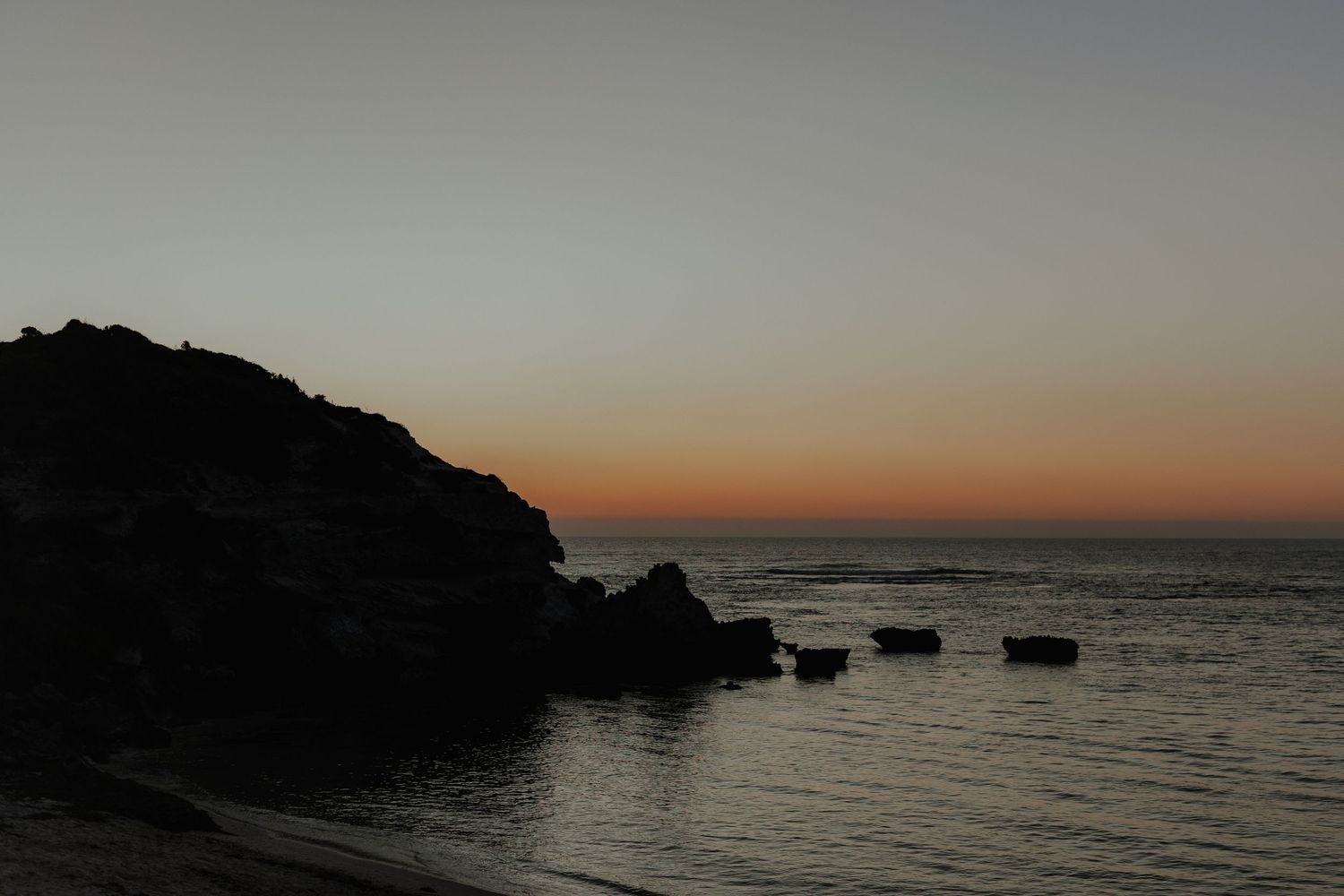 Scenic coastal sunset silhouette with rocky headland and calm ocean waters in warm tones.