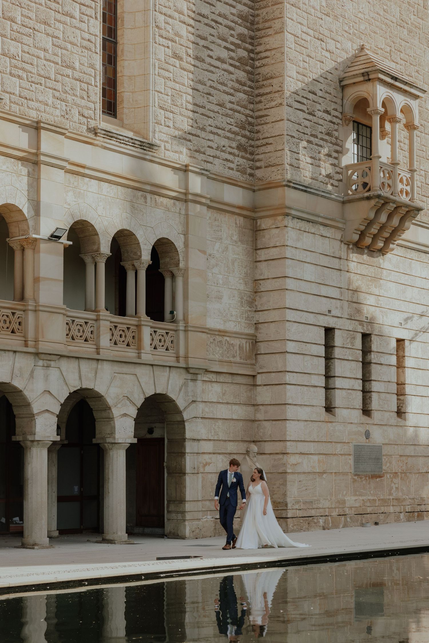 Bride and groom walking beside a reflecting pool in front of a grand stone building with arched colonnades.
