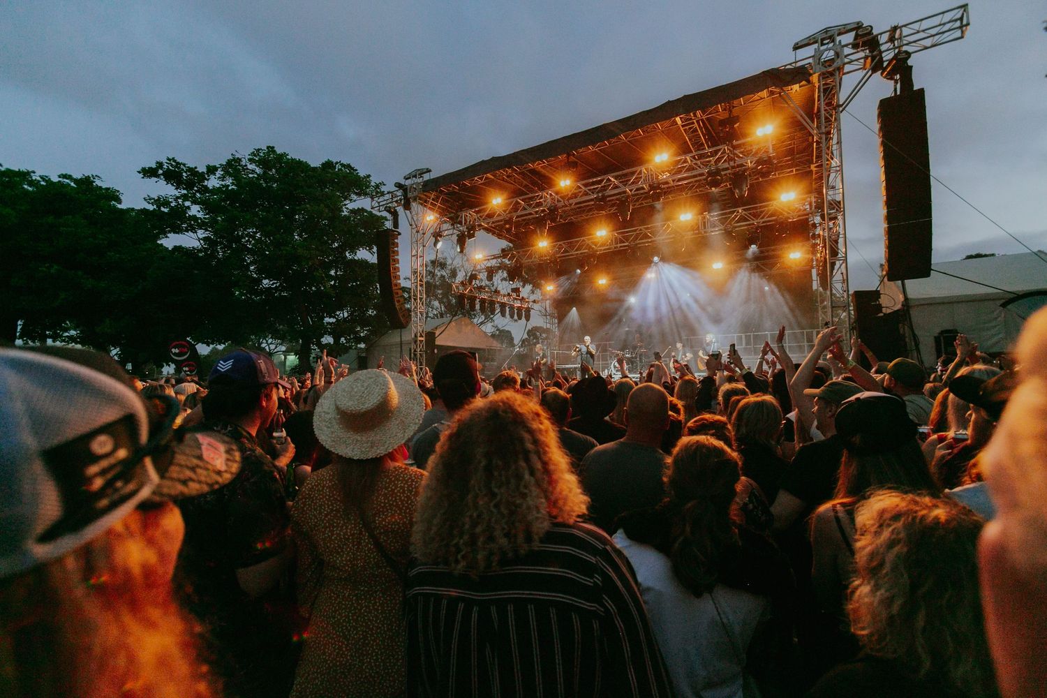 Crowd enjoying an outdoor music festival at dusk with a brightly lit stage and string lights.