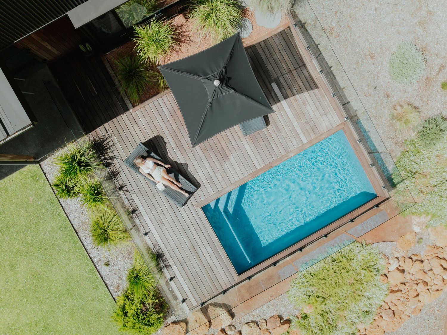 Aerial view of a person relaxing on a timber pool deck beside a small plunge pool with a dark umbrella.