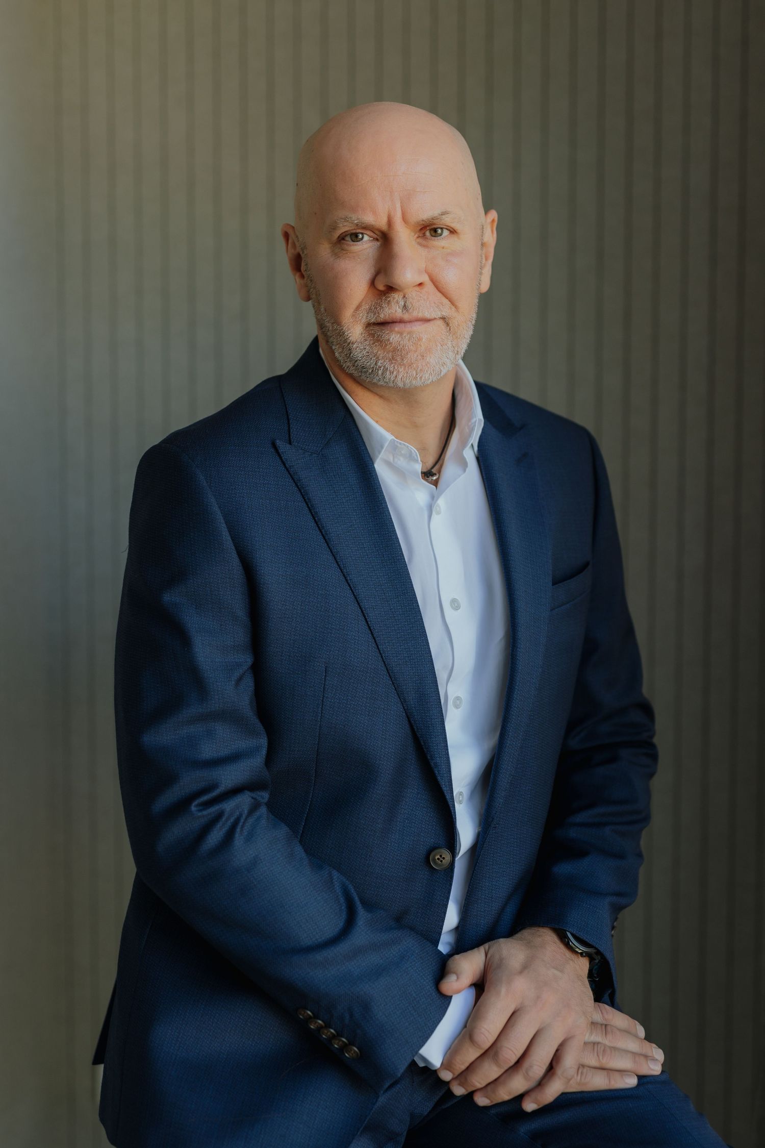 Confident bald man in navy blue suit seated with hands folded, posing for a professional portrait.